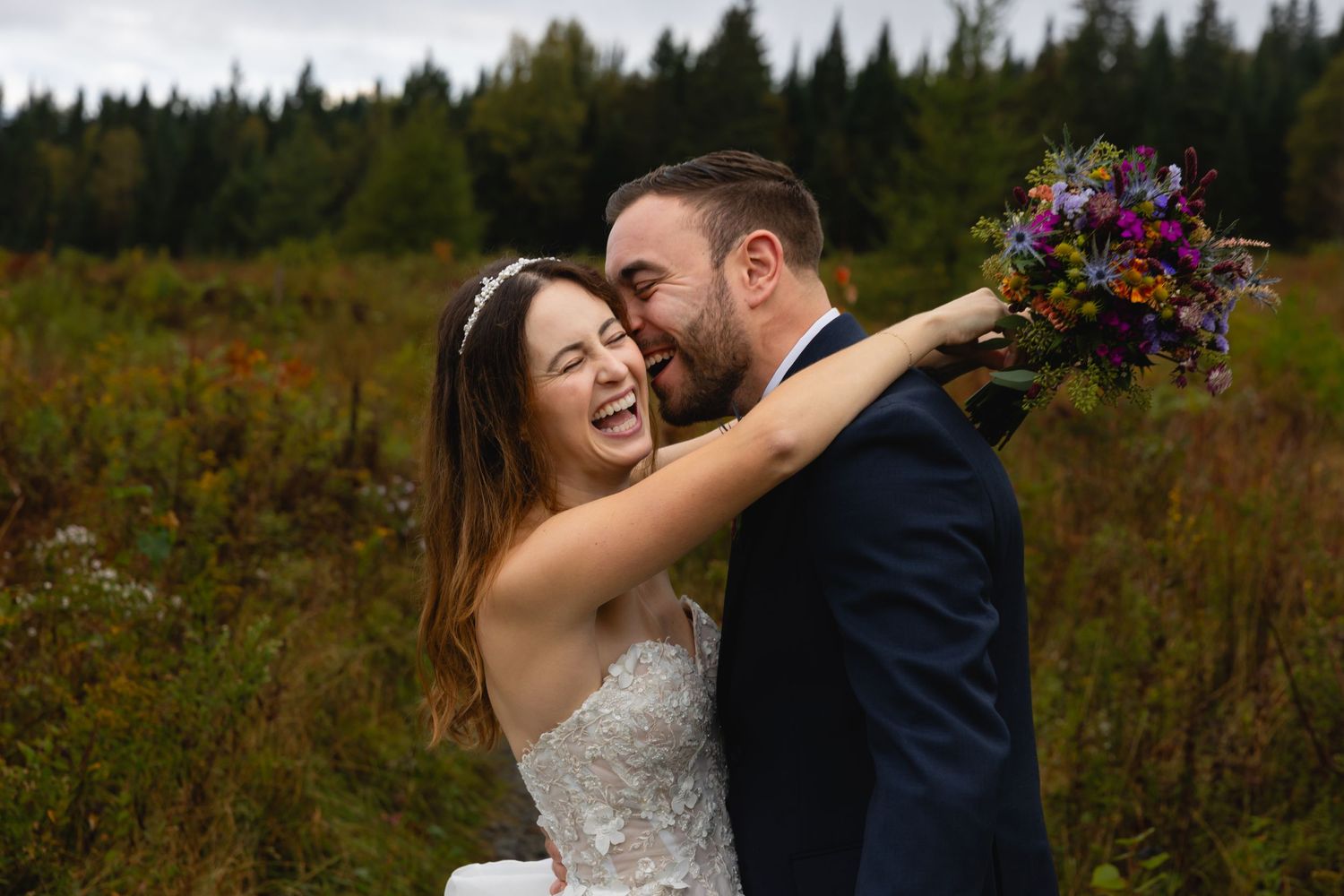 A couple shares a joyful embrace while holding a vibrant wildflower bouquet against mountain scenery.