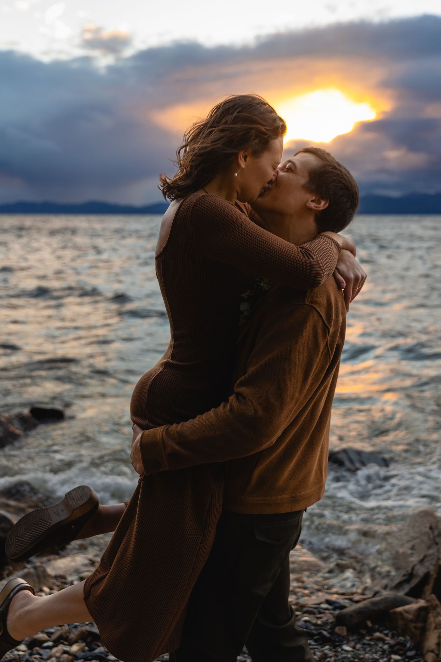 A romantic couple shares a tender kiss on a rocky beach at sunset with dramatic clouds and water in the background.