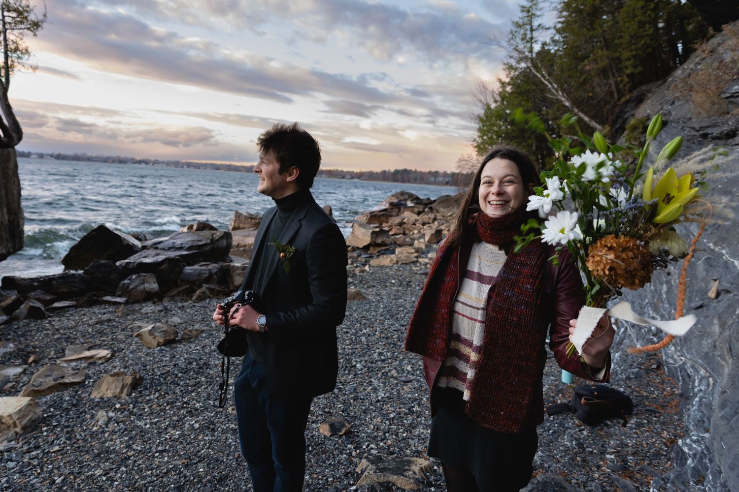 Two people stand on a rocky shoreline at dusk holding camera and flowers against a dramatic cloudy sky.