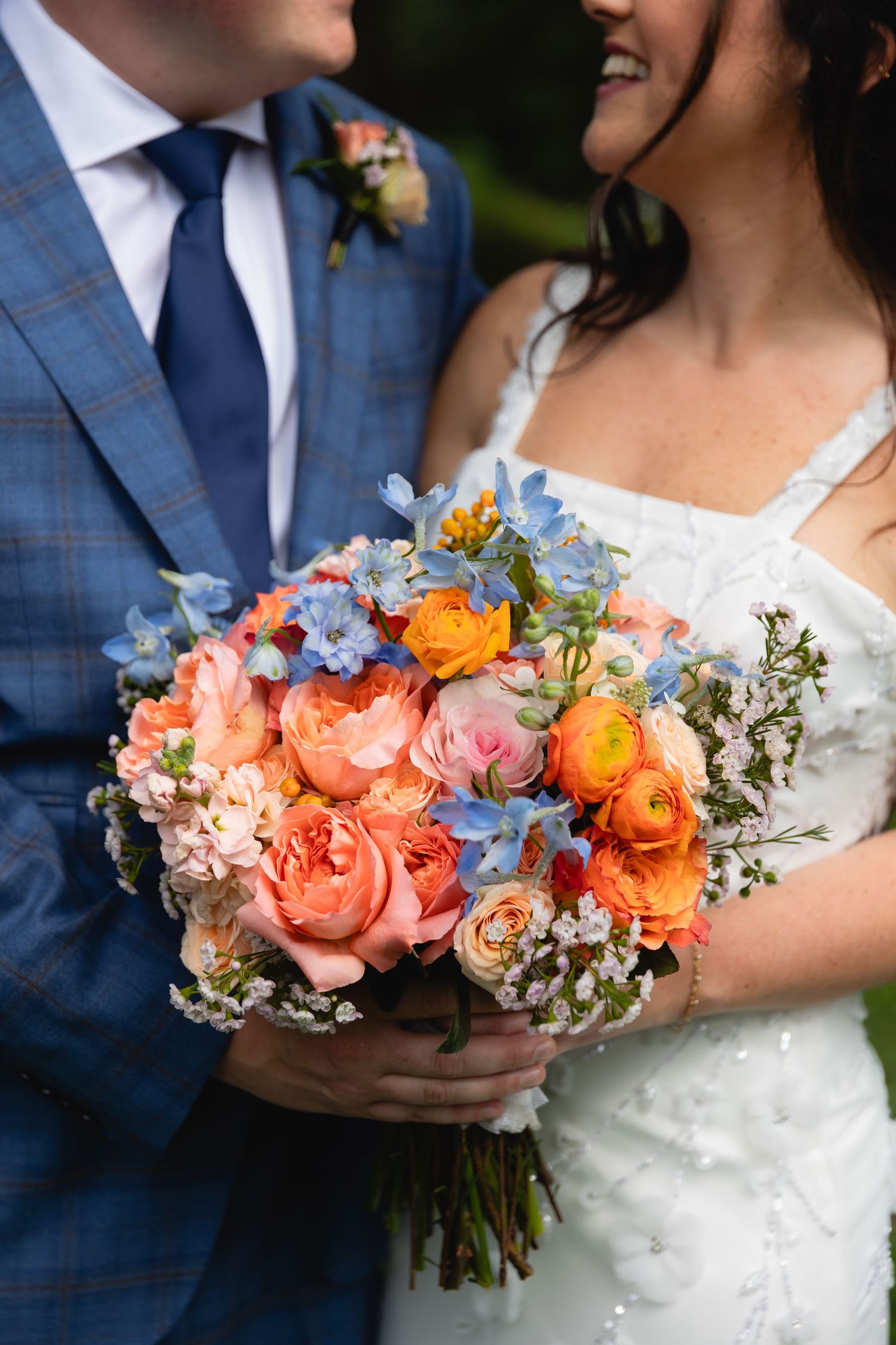 Close-up of a vibrant wedding bouquet with coral roses, orange ranunculus and blue delphinium against formal attire.
