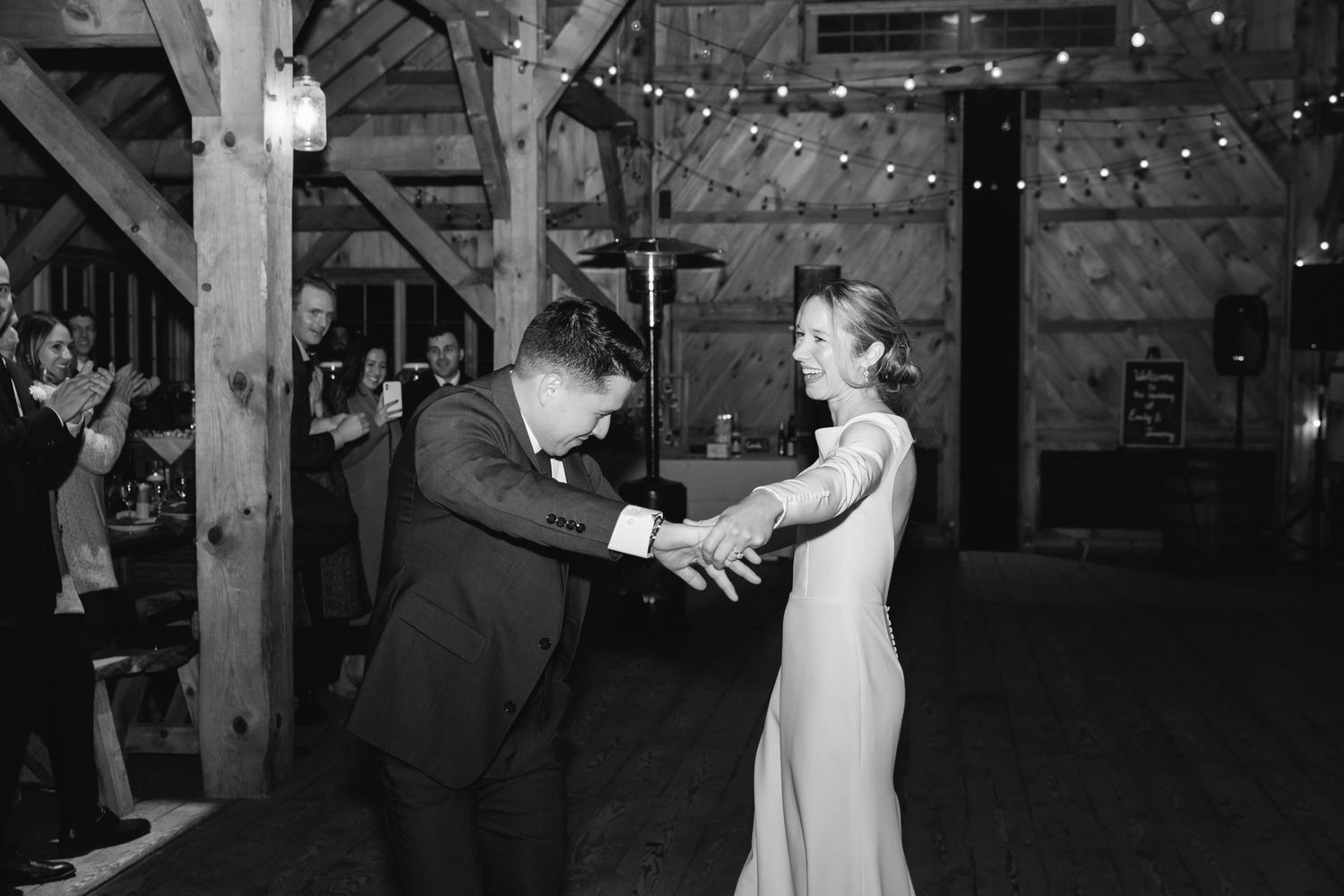 A couple shares their first dance at a rustic barn wedding reception illuminated by string lights.