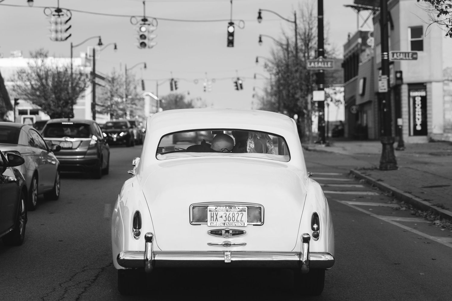A classic white vintage car seen from behind on a city street with traffic lights and buildings in black and white.