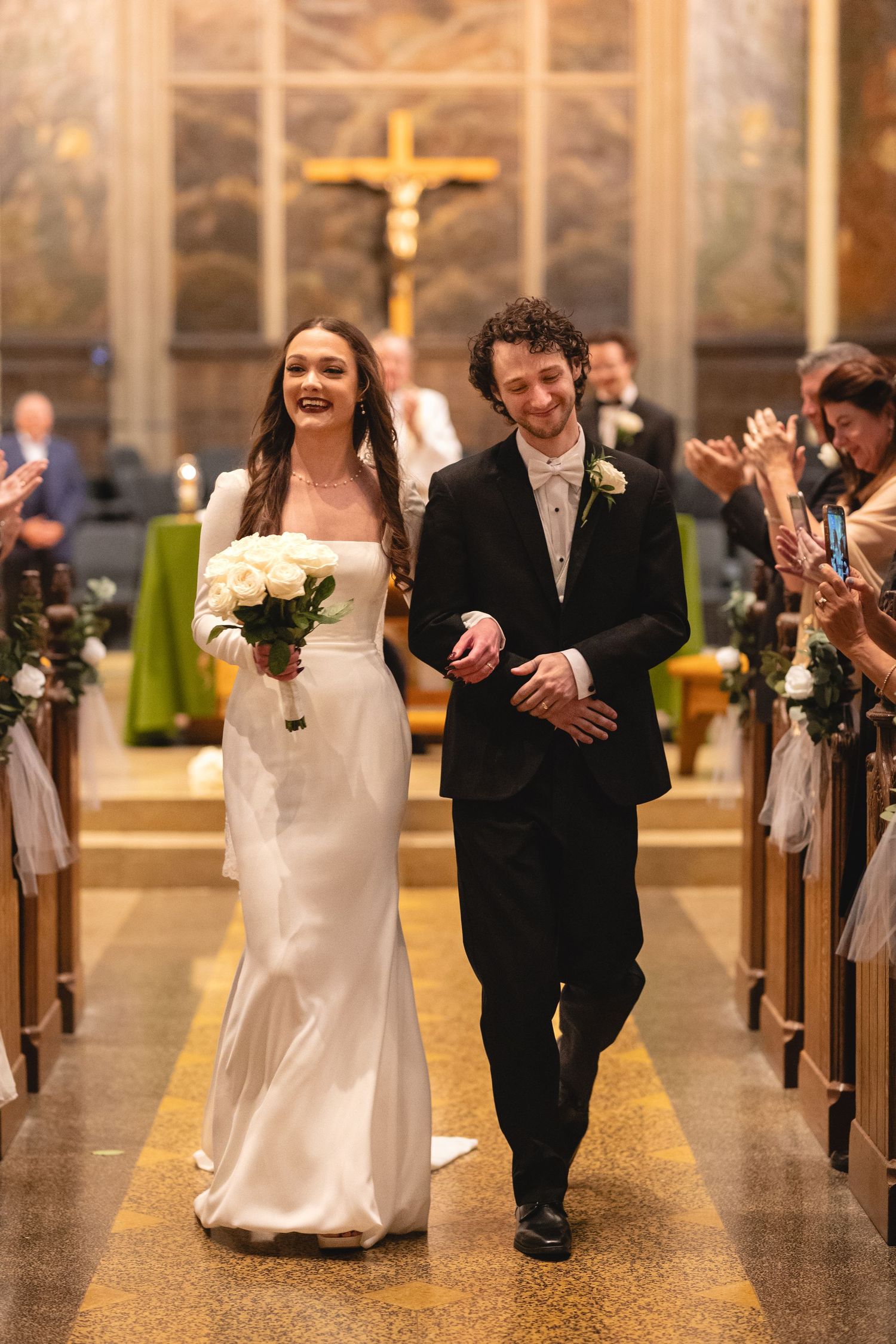 Newlyweds walk down the church aisle while guests applaud, with a crucifix and religious artwork visible in the background.