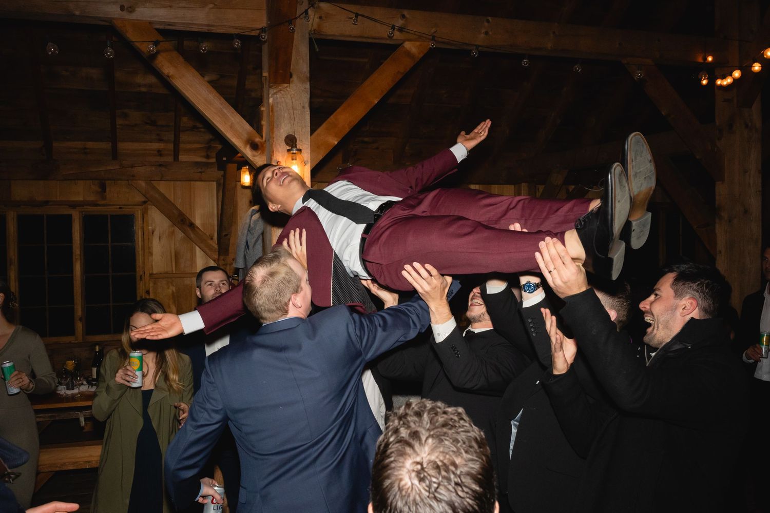 A person in a burgundy suit is being lifted up by a crowd at what appears to be a party in a rustic venue.