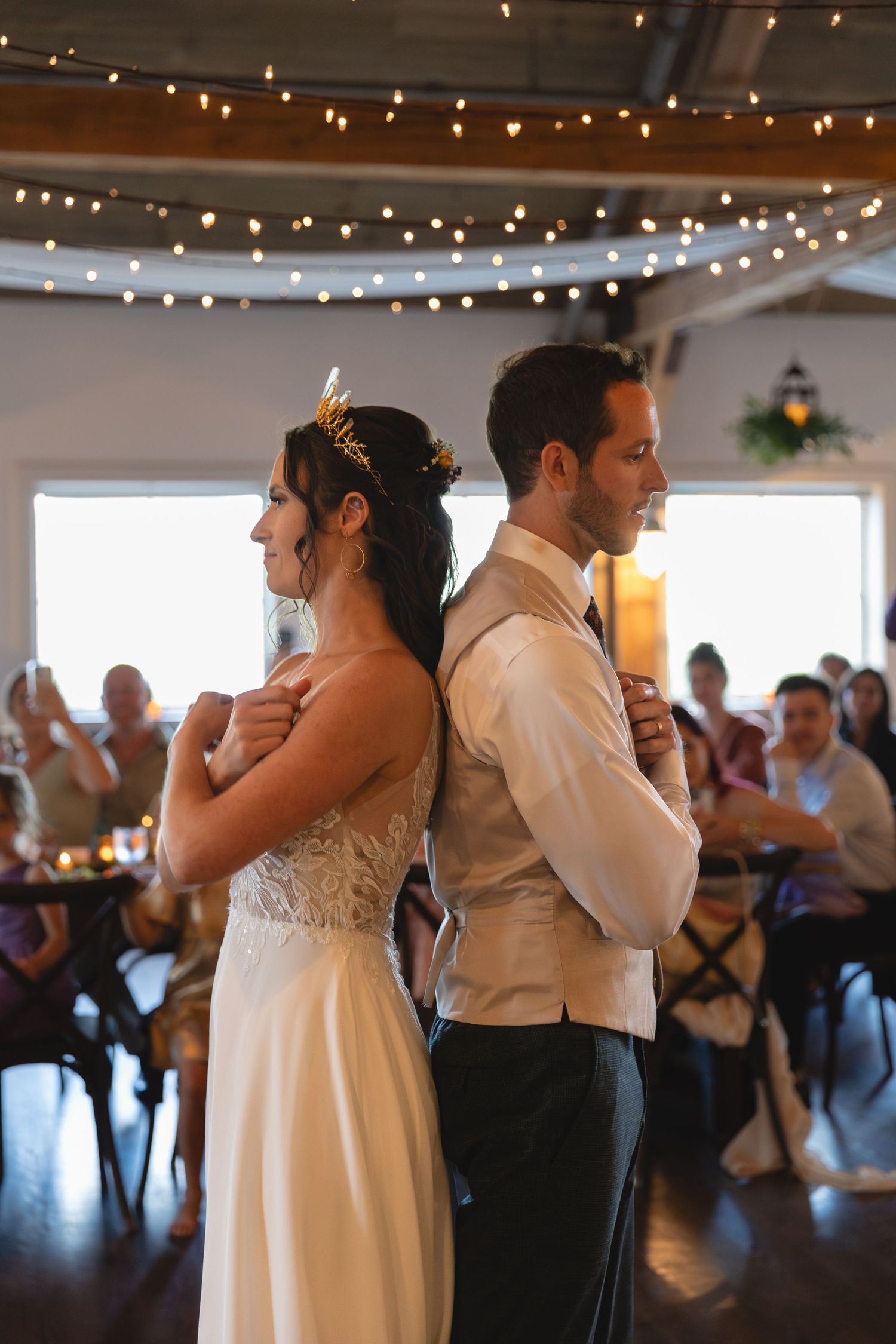 Couple in formal attire standing back to back under string lights at indoor venue.