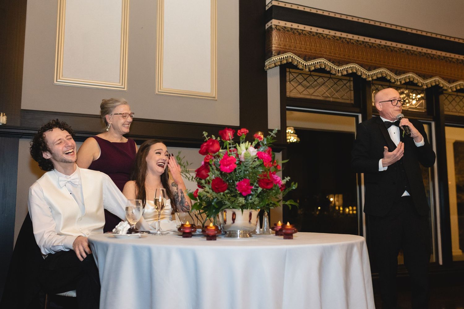 People at a wedding reception table laugh while listening to a speaker in formal attire delivering a toast.