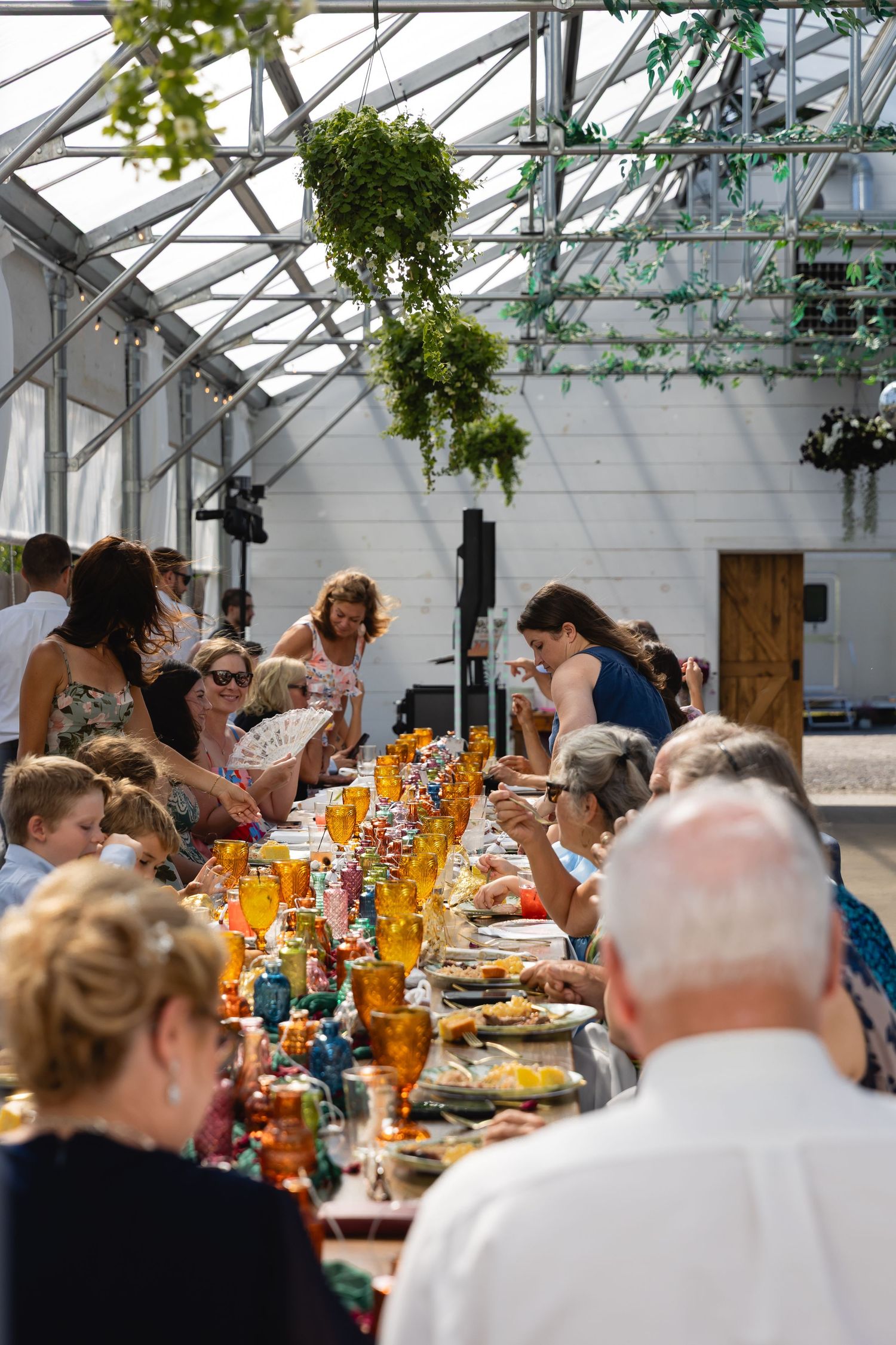 Guests gather around long tables decorated with orange flowers in a bright greenhouse venue for a special celebration.