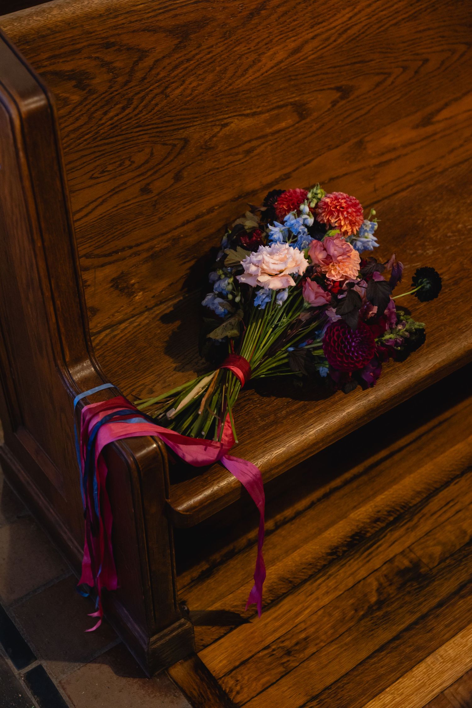 A vibrant wedding bouquet with pink and red flowers tied with purple ribbons rests on a wooden church pew.