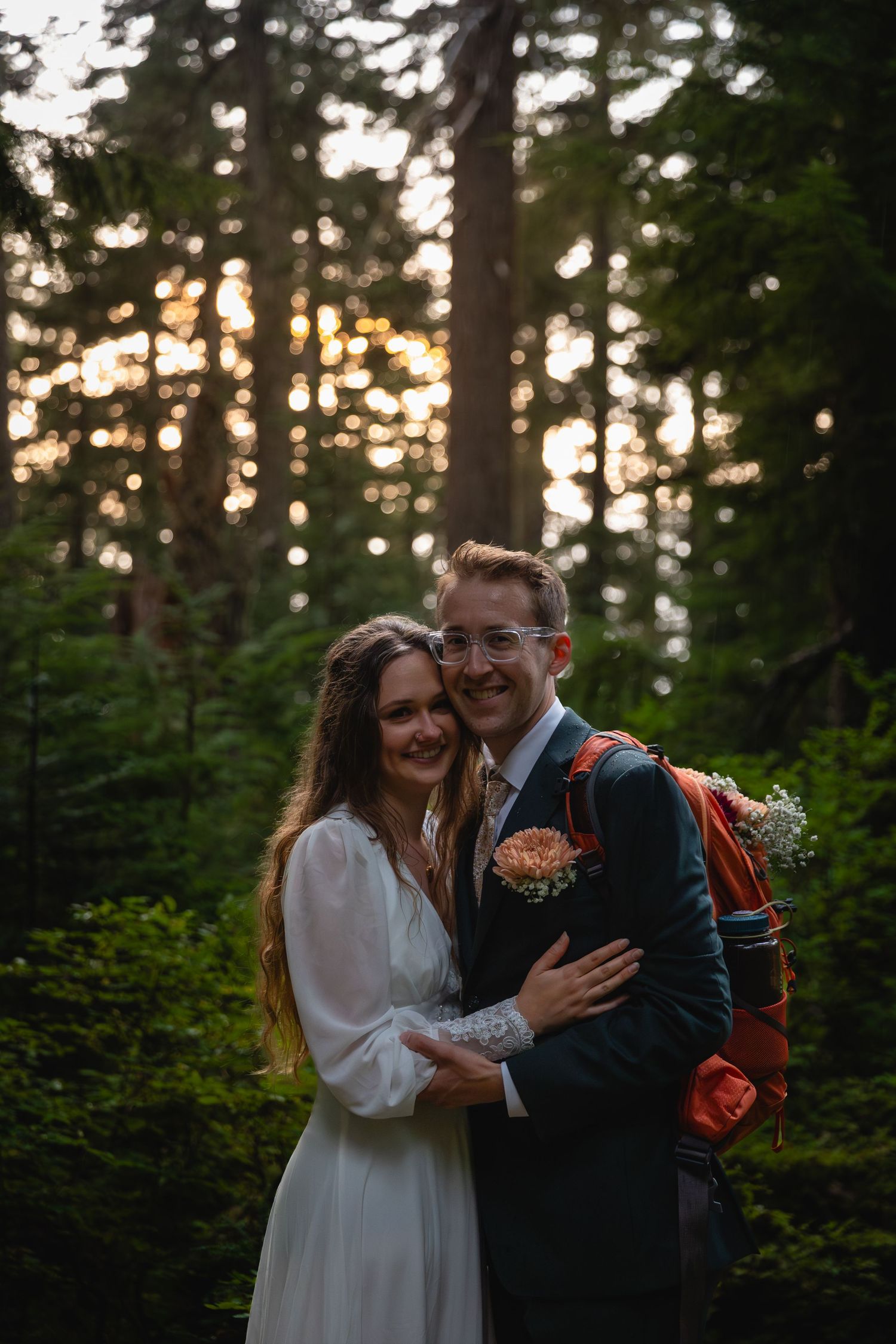 A couple shares a joyful embrace while standing in a lush evergreen forest at sunset.