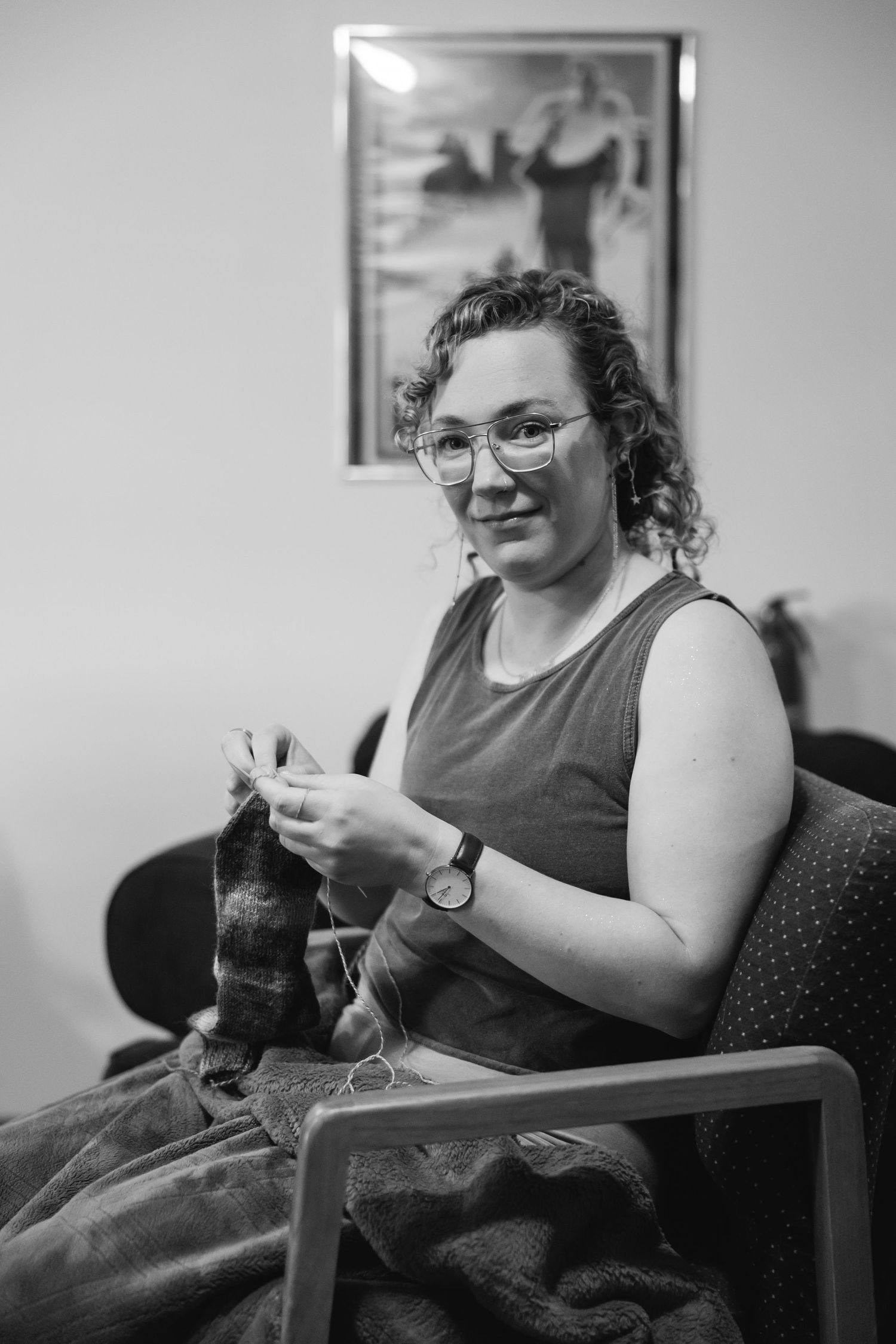 A person sits in a chair and knits while wearing glasses and a dark sleeveless top in a black and white photo series.