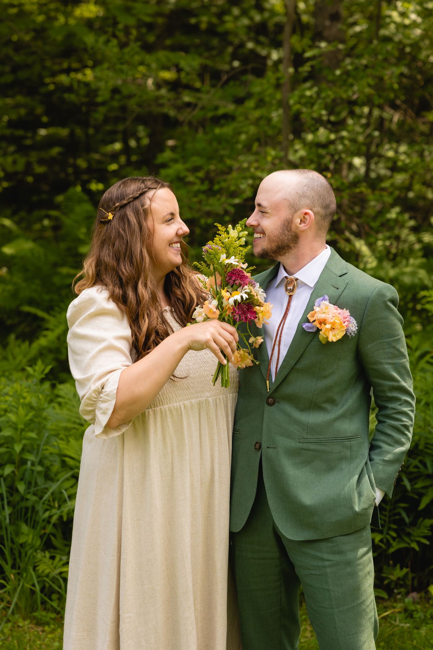 A couple shares a joyful moment in a forest setting, with the bride in cream and groom in forest green attire.