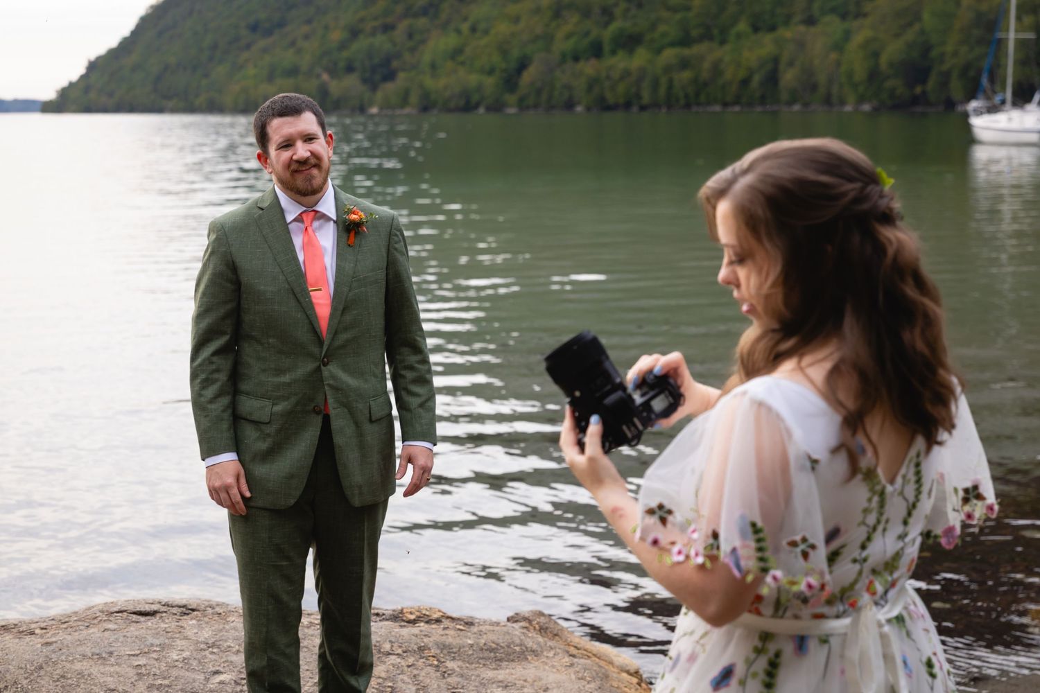 A photographer captures a wedding scene by a scenic lake with mountains in the background.