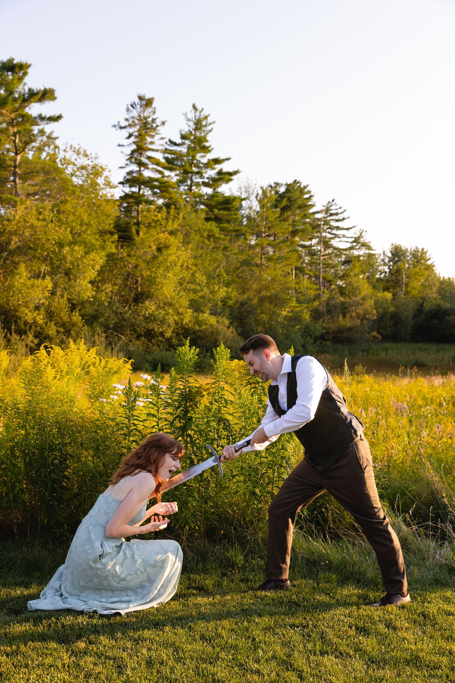 A couple in formal attire playfully interact in a golden meadow with evergreen trees in the background at sunset.