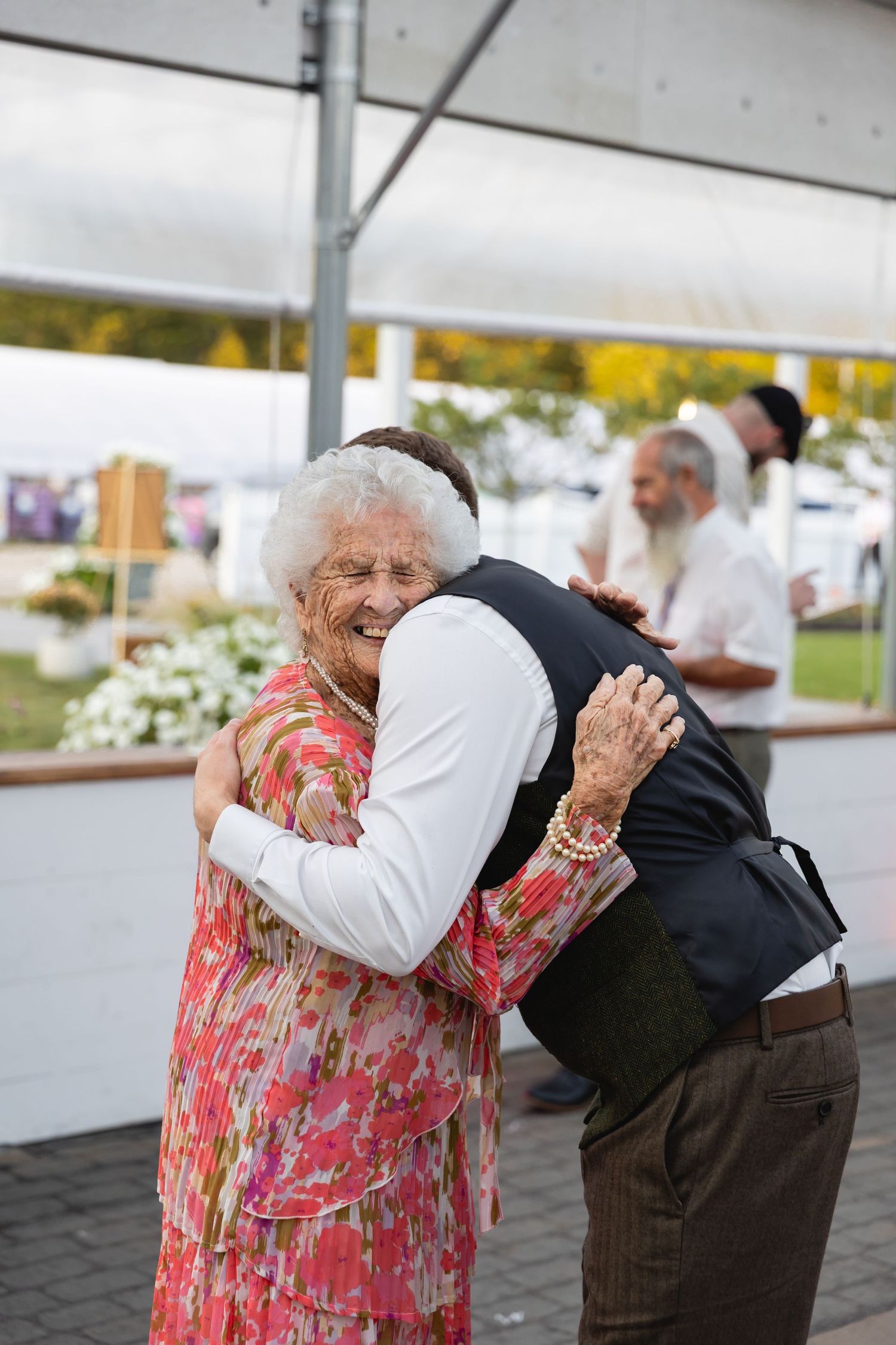 Two elderly people share an emotional embrace at an outdoor event under a white tent.