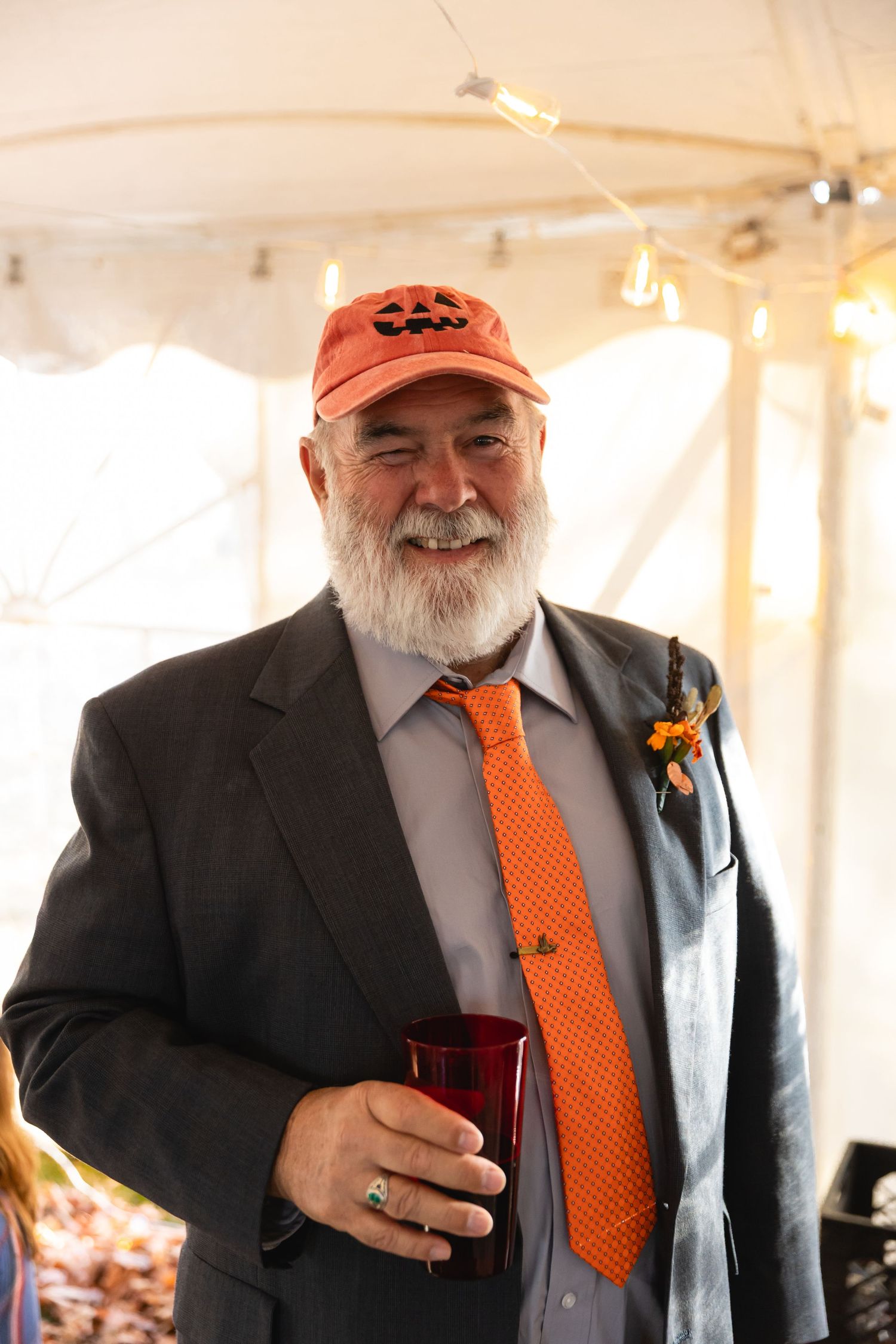 A person wearing a black suit jacket and orange tie holds a dark beverage while smiling in a tent setting.