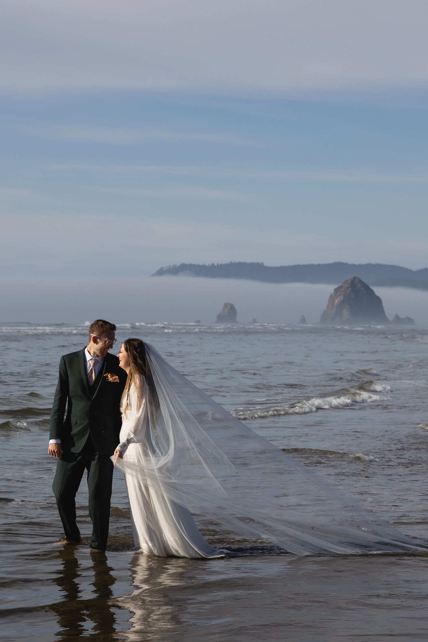 A couple stands in shallow ocean waves during a romantic beach wedding photoshoot with a dramatic coastal backdrop.
