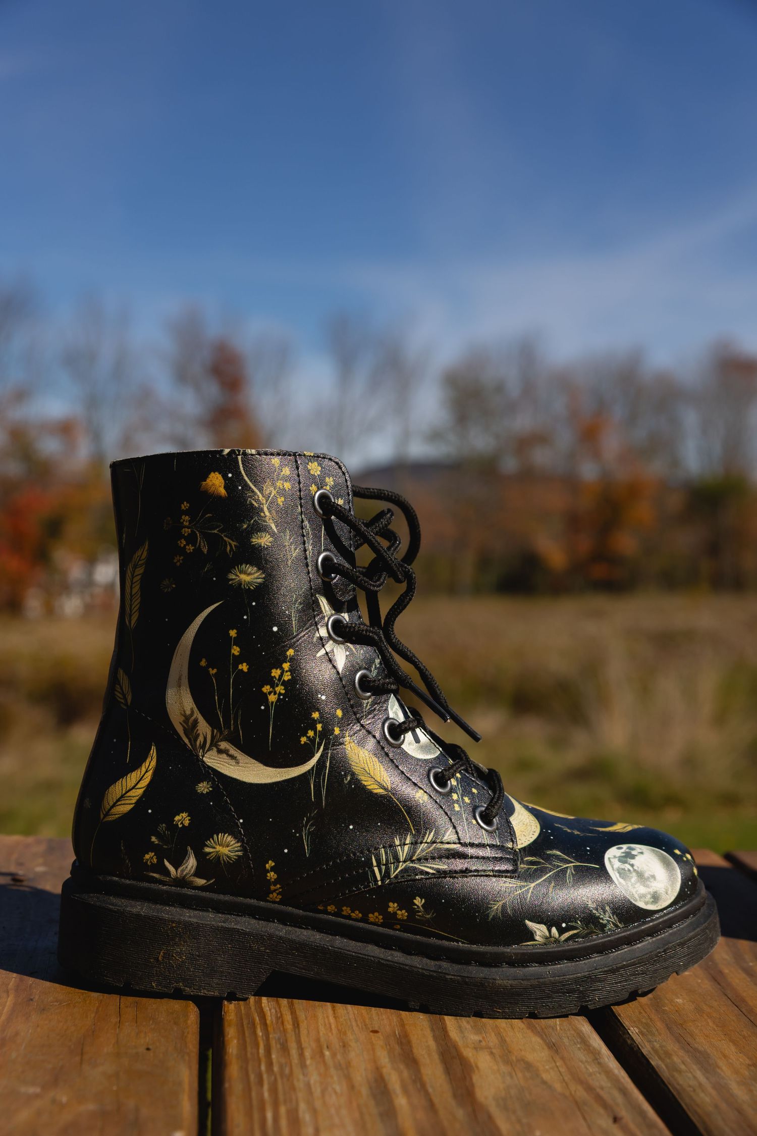 Hand-painted black leather boots with celestial moon and star designs displayed on wooden surface in autumn outdoor setting.