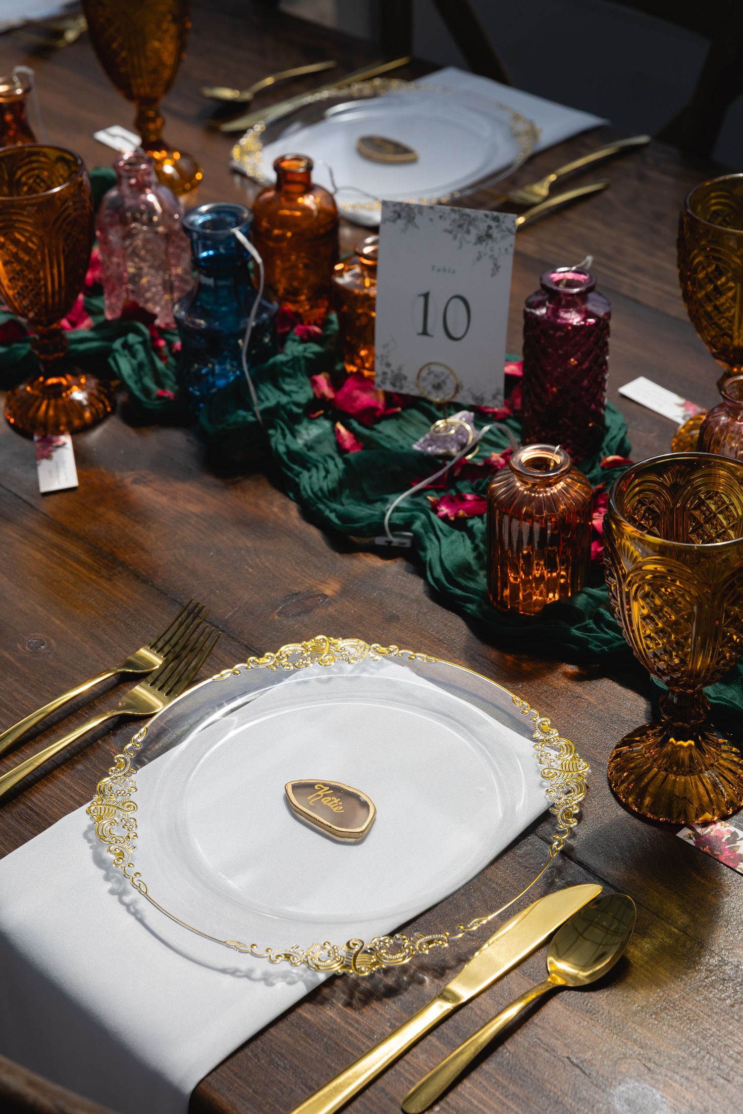 Elegant table setting with gold cutlery, white plates with gold trim, and colorful vintage glass bottles on green velvet runner.