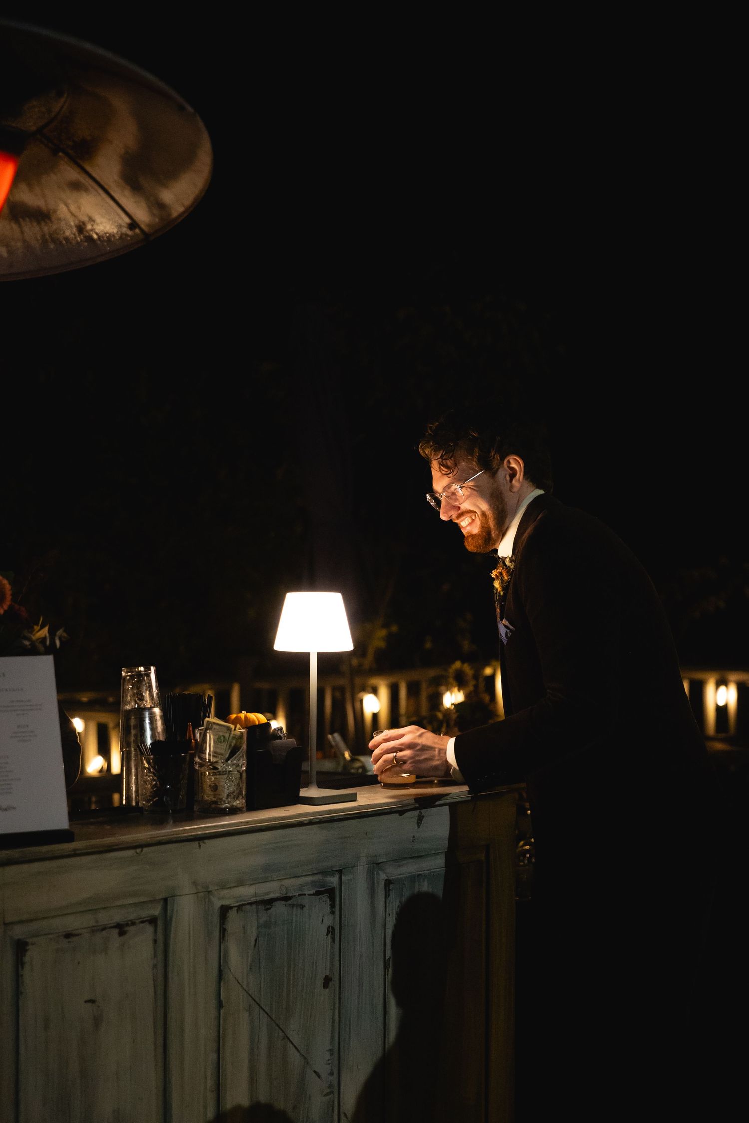 Groom working at a rustic outdoor bar illuminated by a small lamp at night.
