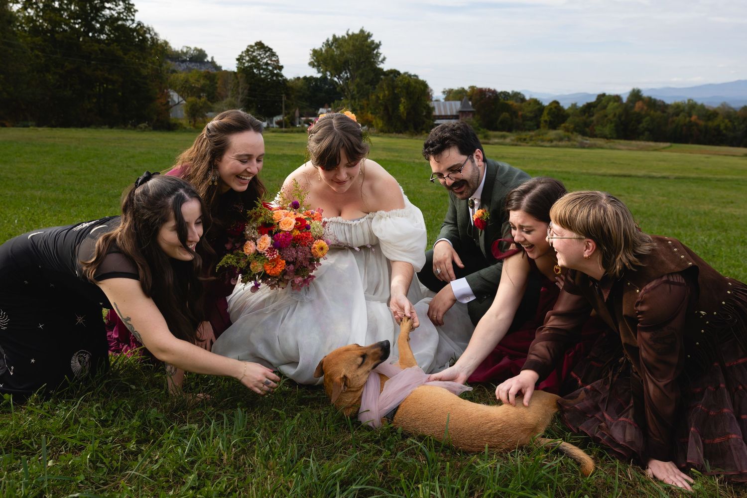 Wedding party gathered on grass petting a dog, bride holding colorful autumn bouquet.