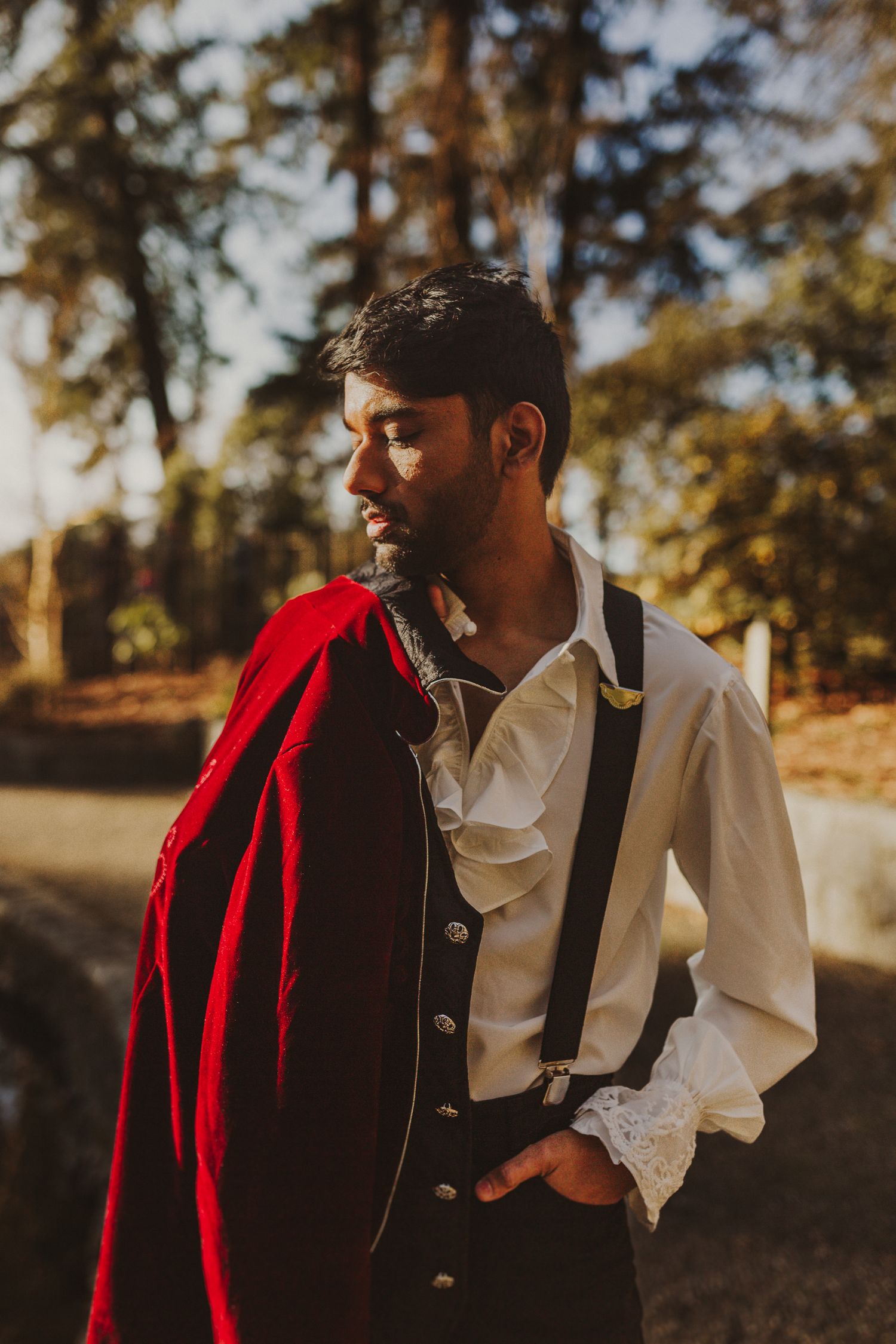 A bearded man in a white ruffled shirt and suspenders holds a red cape outdoors in golden sunlight.