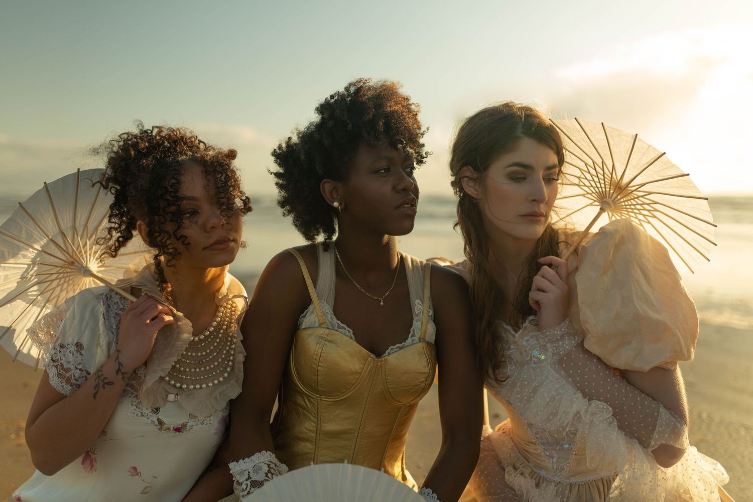 Three women in vintage lace and satin gowns holding parasols on sunlit beach.