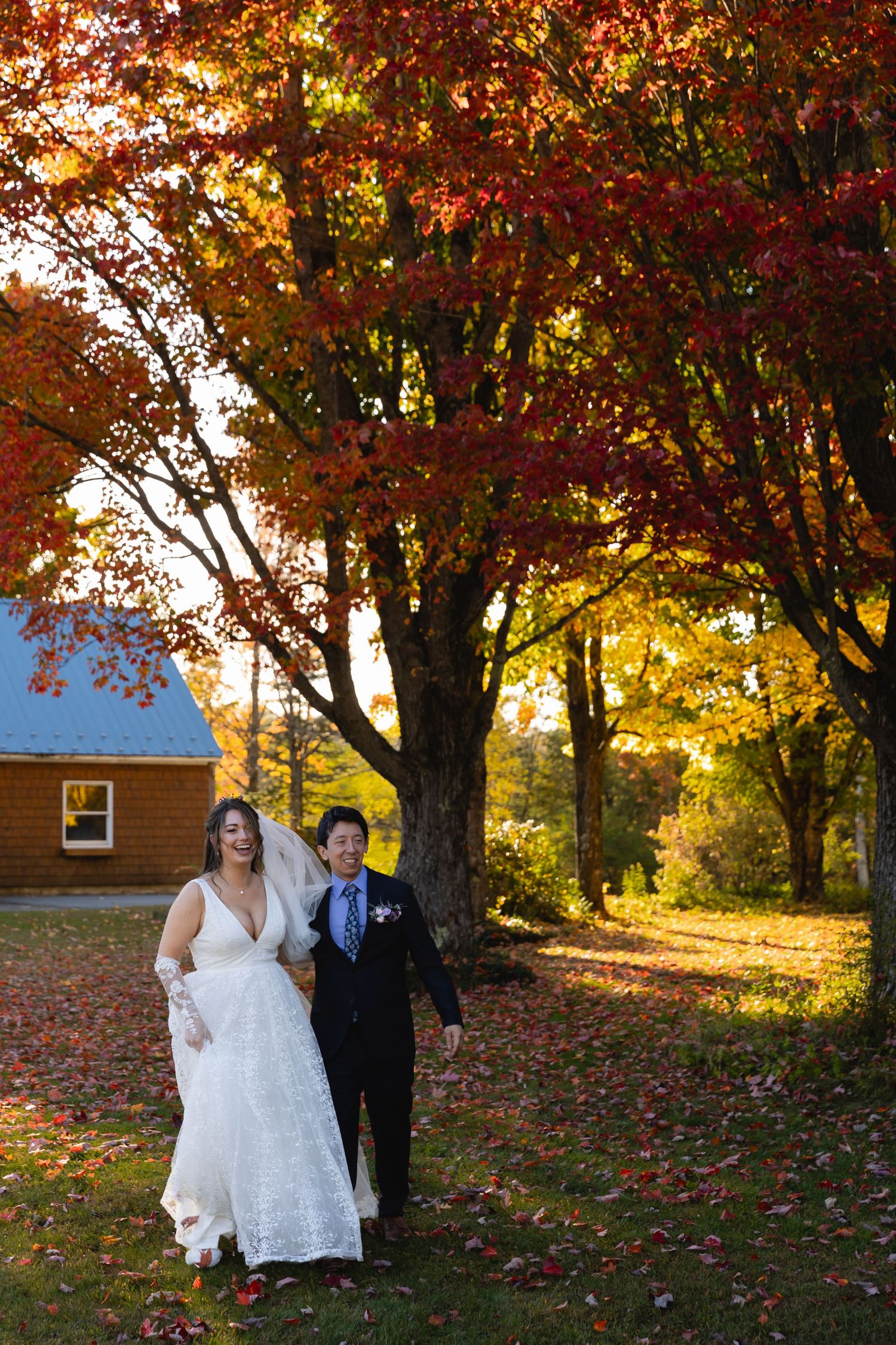 Bride and groom walk arm in arm beneath vibrant red and orange autumn maple trees.