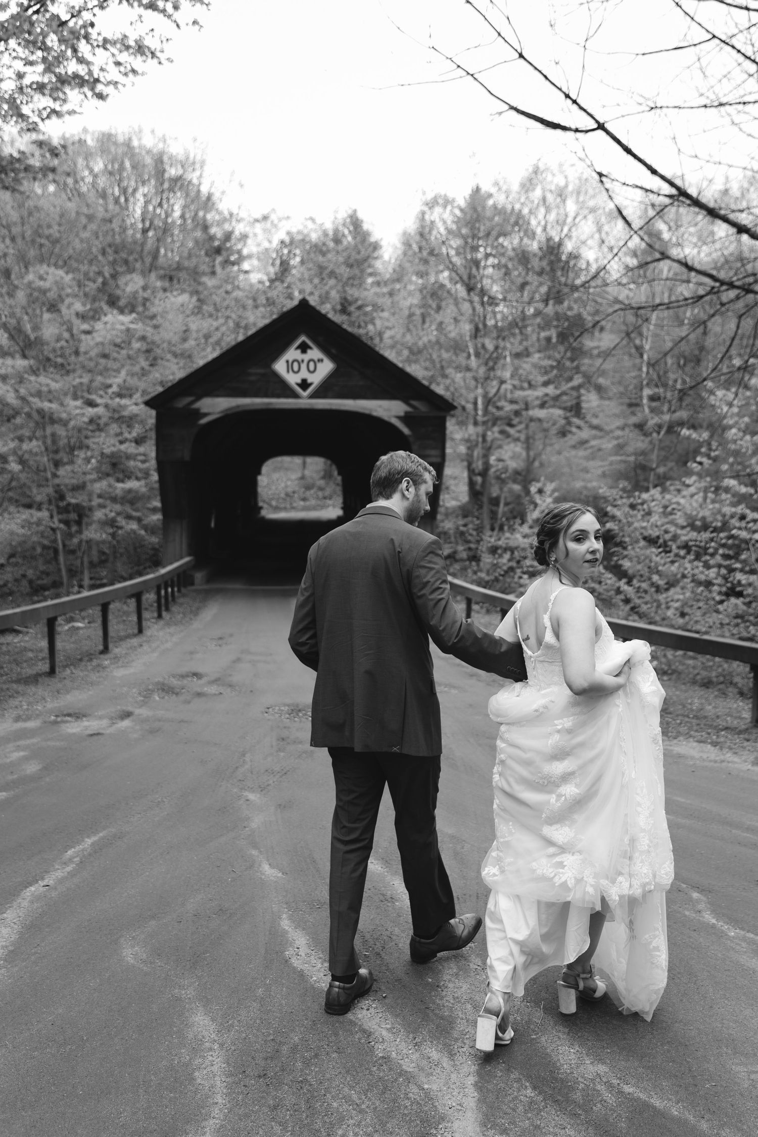 Black and white photo of bride and groom walking toward a covered bridge surrounded by autumn trees.
