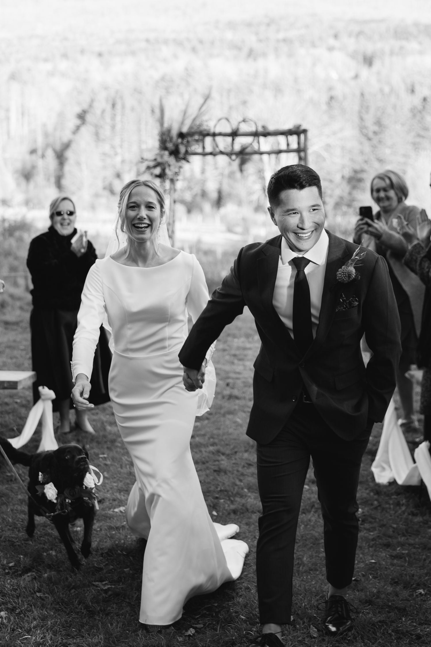 A joyful newlywed couple runs hand-in-hand through a garden with a black dog following behind in a black and white photo.