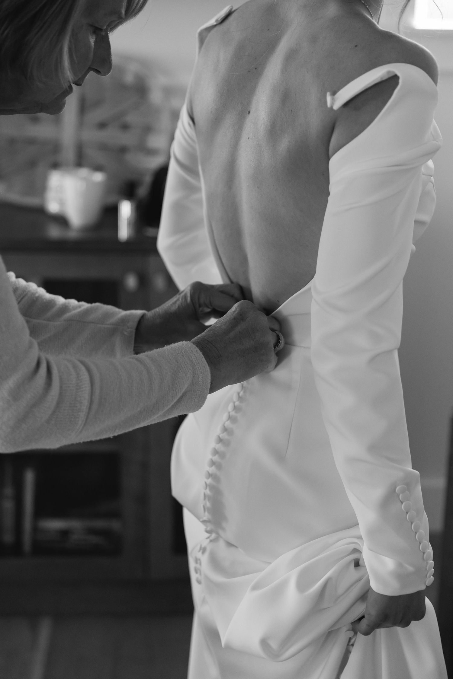 Black and white photo of someone helping button up an elegant white wedding dress with delicate details.