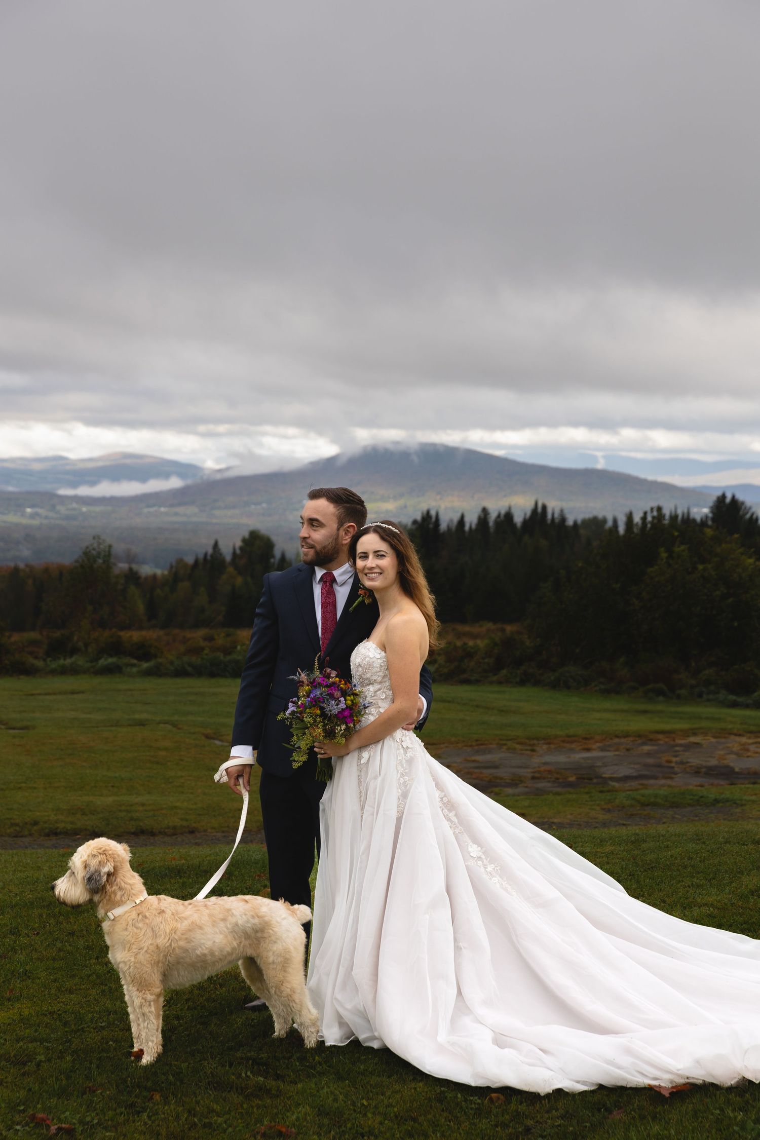 A wedding couple poses with their white dog against a dramatic mountain backdrop.
