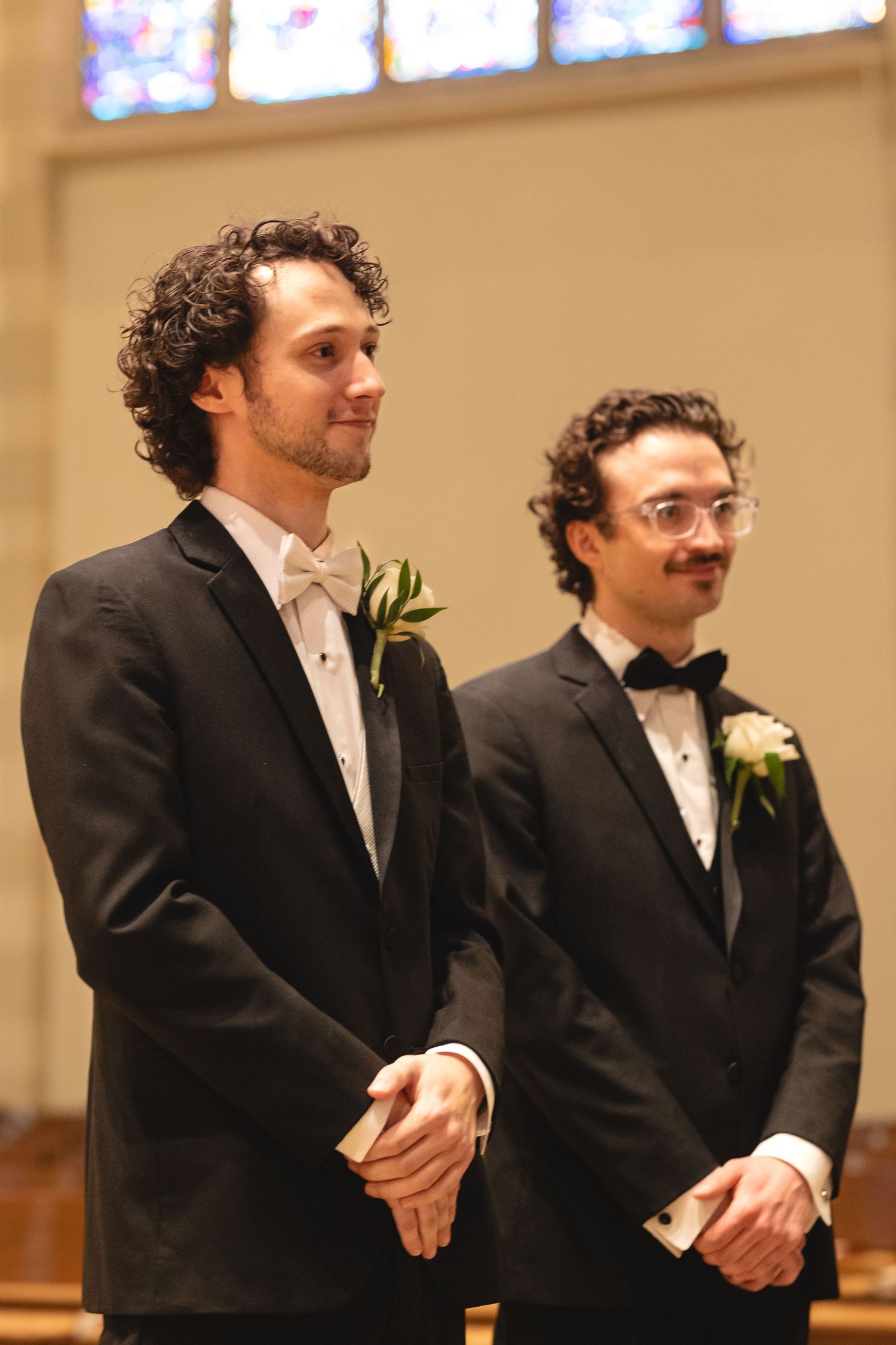 Two groomsmen standing at altar in black tuxedos with white boutonnieres and bow ties at formal wedding ceremony.