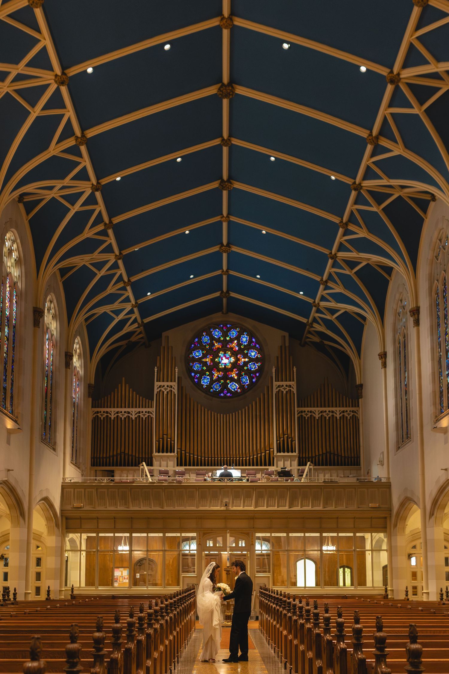 A couple stands in the aisle of a beautiful church with vaulted wooden ceilings, stained glass windows and a pipe organ.