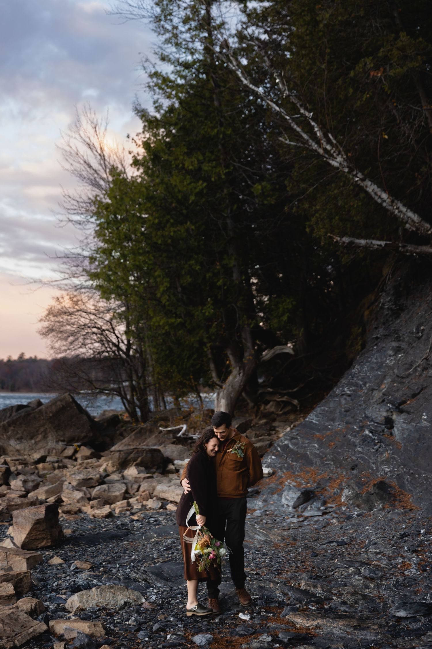 Two bundled-up figures explore a rocky winter shoreline at sunset with tall trees and moody clouds in the background.