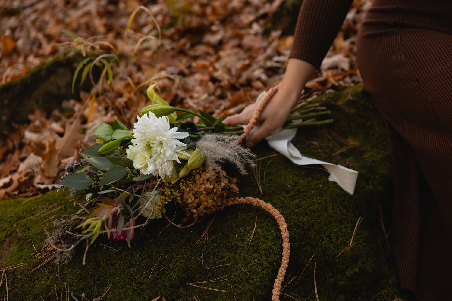 Hand placing white chrysanthemum flower bouquet on mossy log surrounded by autumn leaves in forest setting.