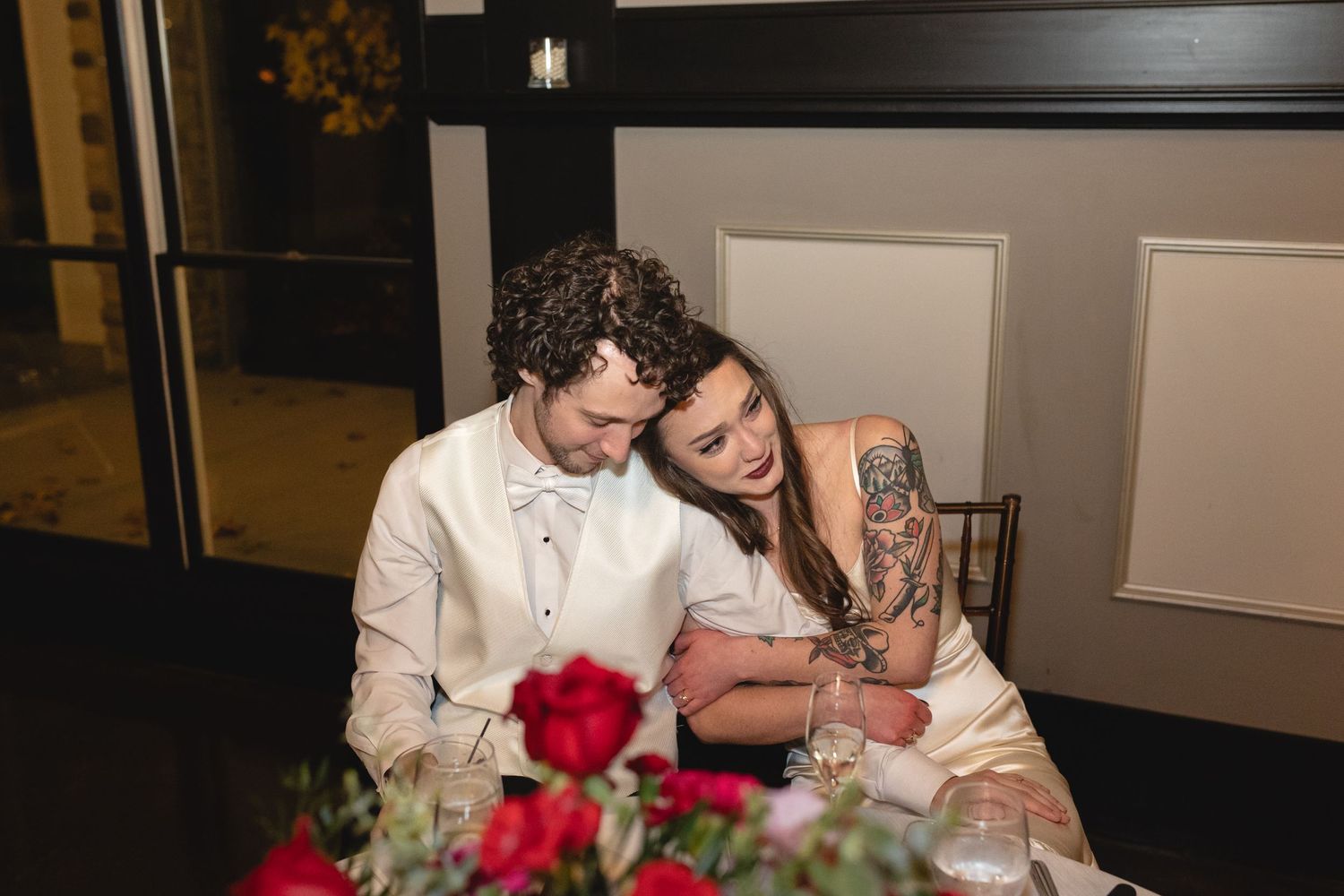A couple shares an intimate moment at their wedding reception with red roses decorating the table.