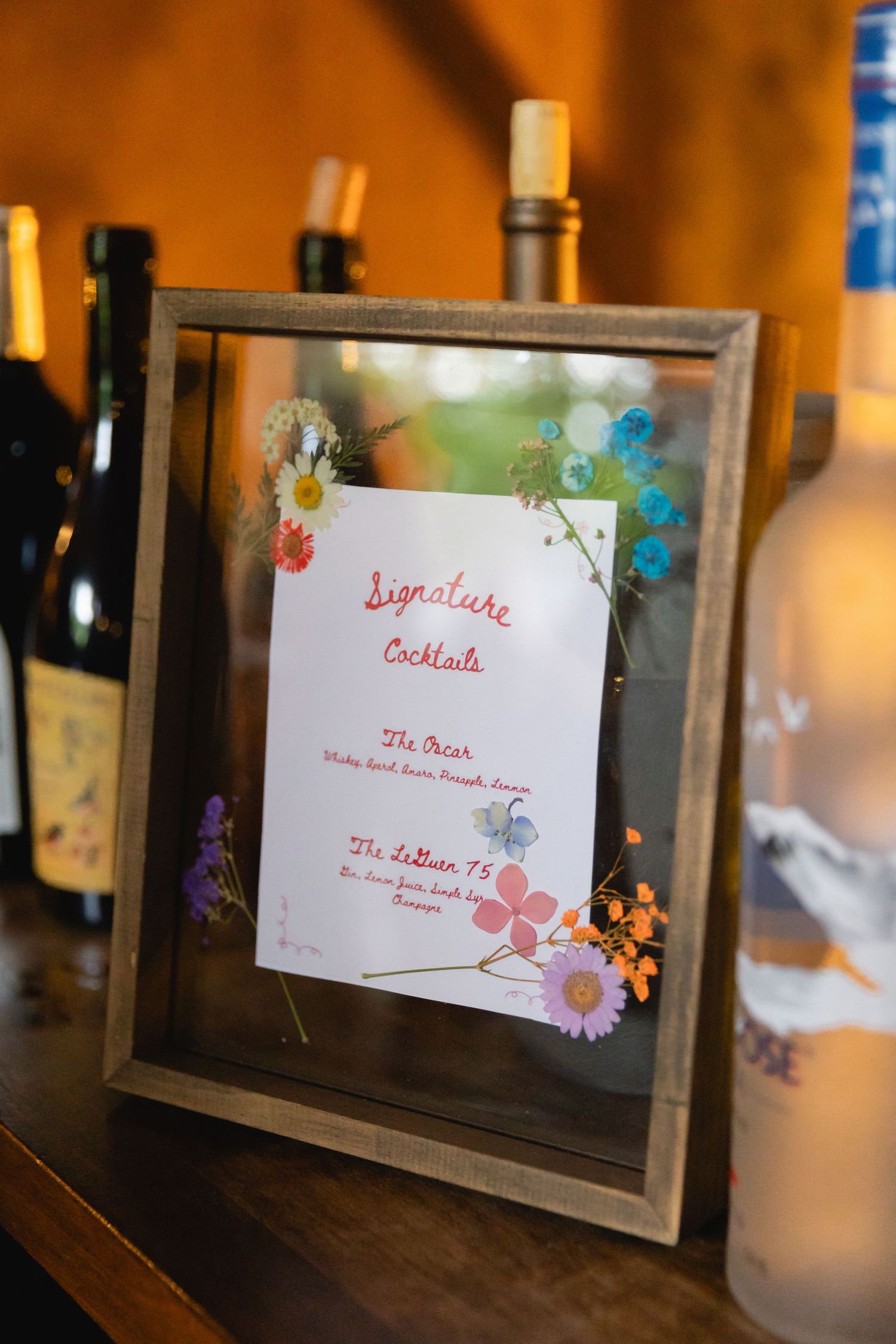 A framed document with floral decorations displayed on a bar counter with wine bottles in the background.