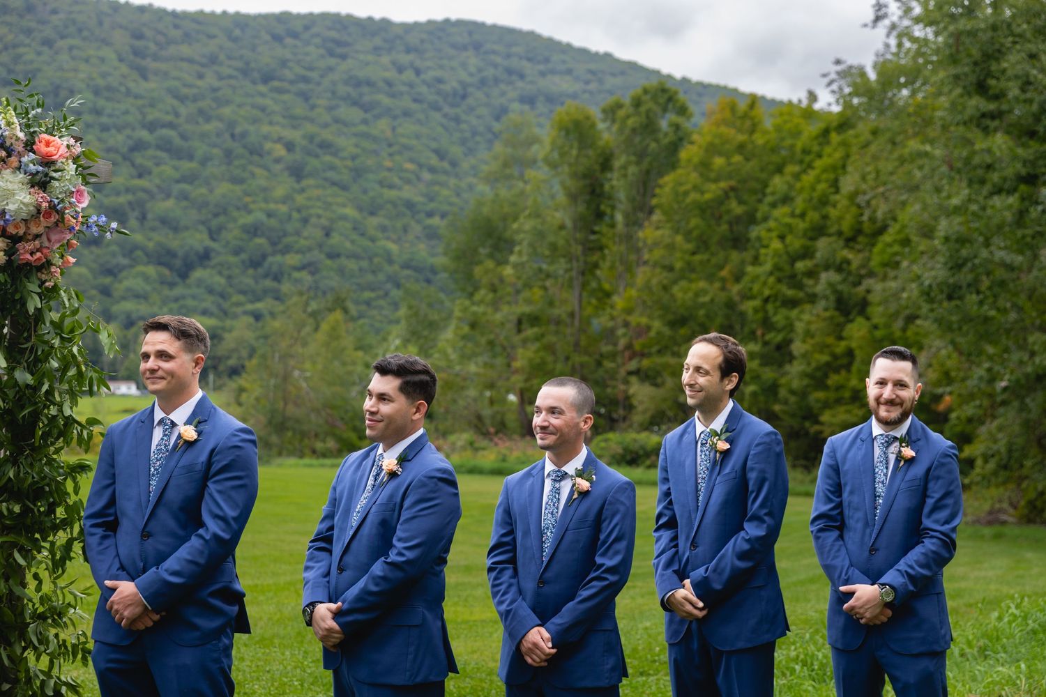 Groomsmen in matching royal blue suits stand in line on a grassy hillside with mountain views in the background.