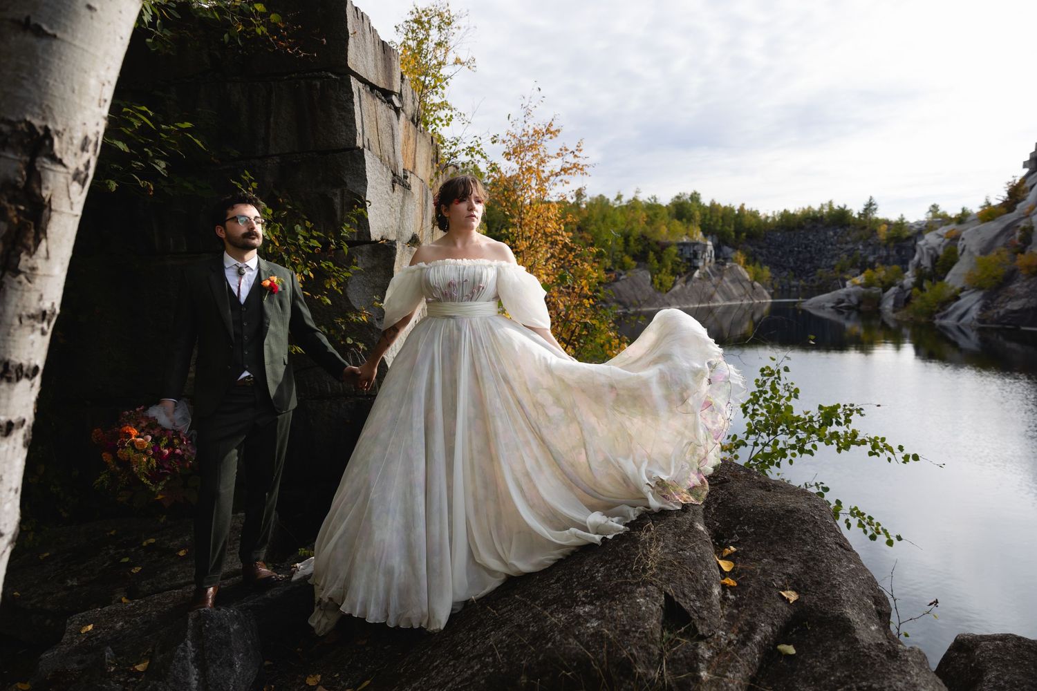 A couple stands on rocky terrain by a lake, with the bride's dress flowing dramatically in the wind.