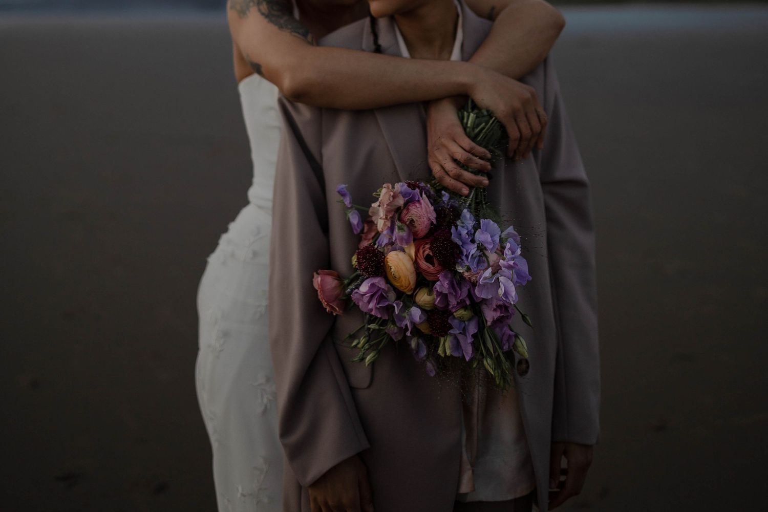 A couple embraces while holding a vibrant purple and pink floral bouquet against moody lighting.