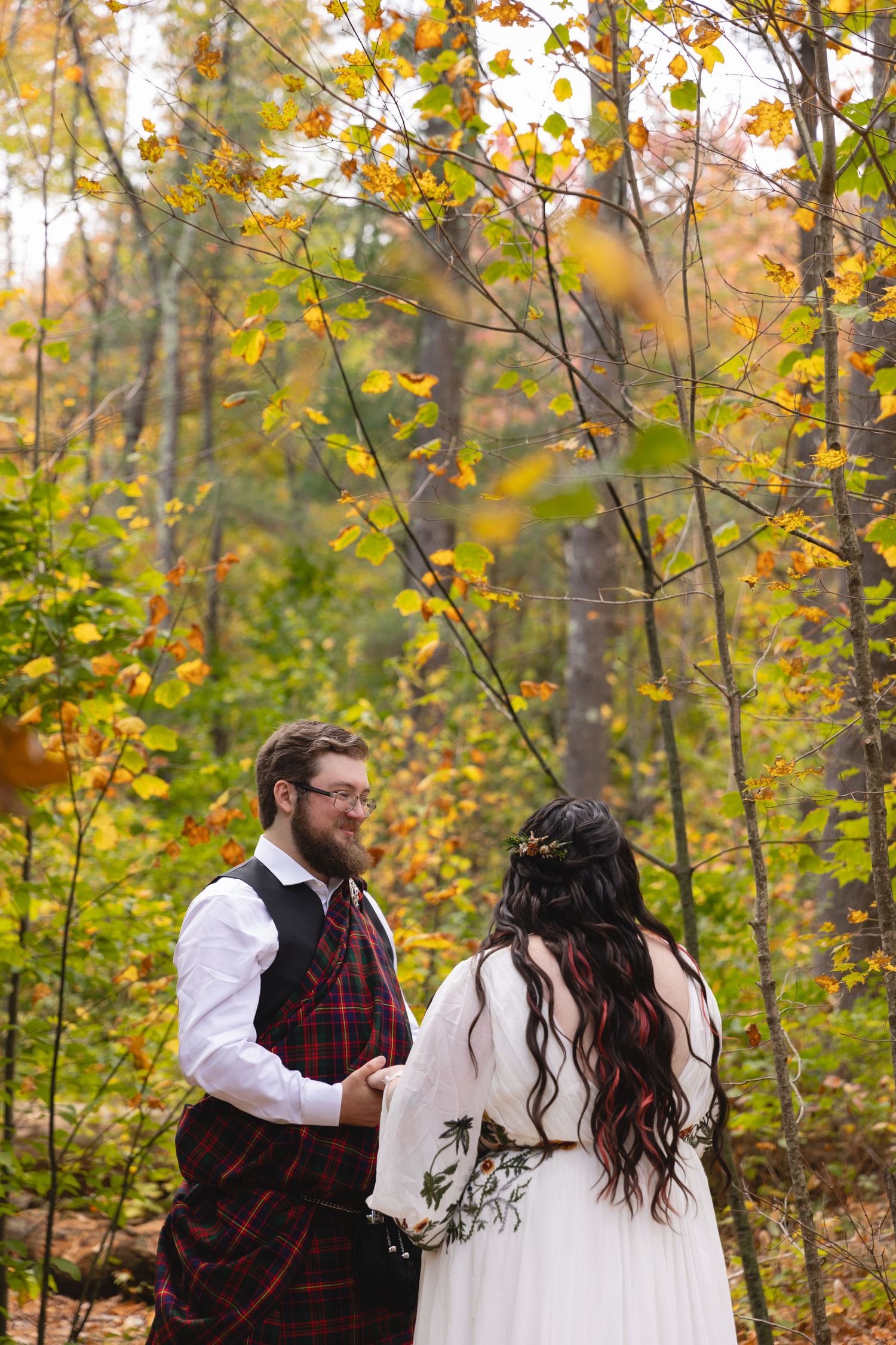 Couple in Scottish wedding attire share intimate moment among fall foliage.