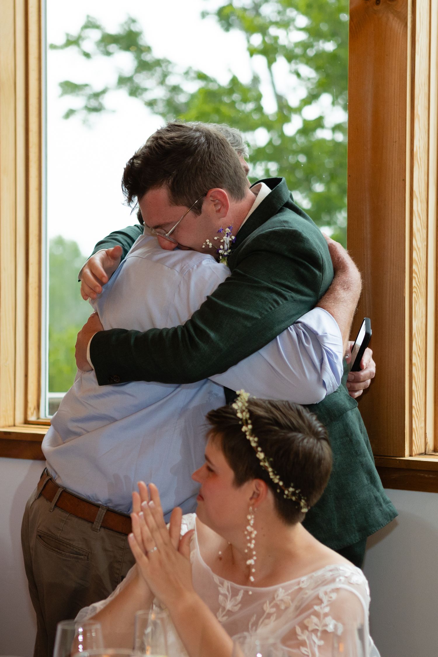 Embracing figures at a wedding reception by sunny windows with decorative floral centerpieces.