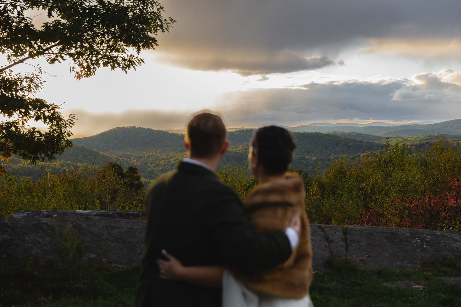 Wedding couple gazes at mountain landscape during golden hour while embracing.