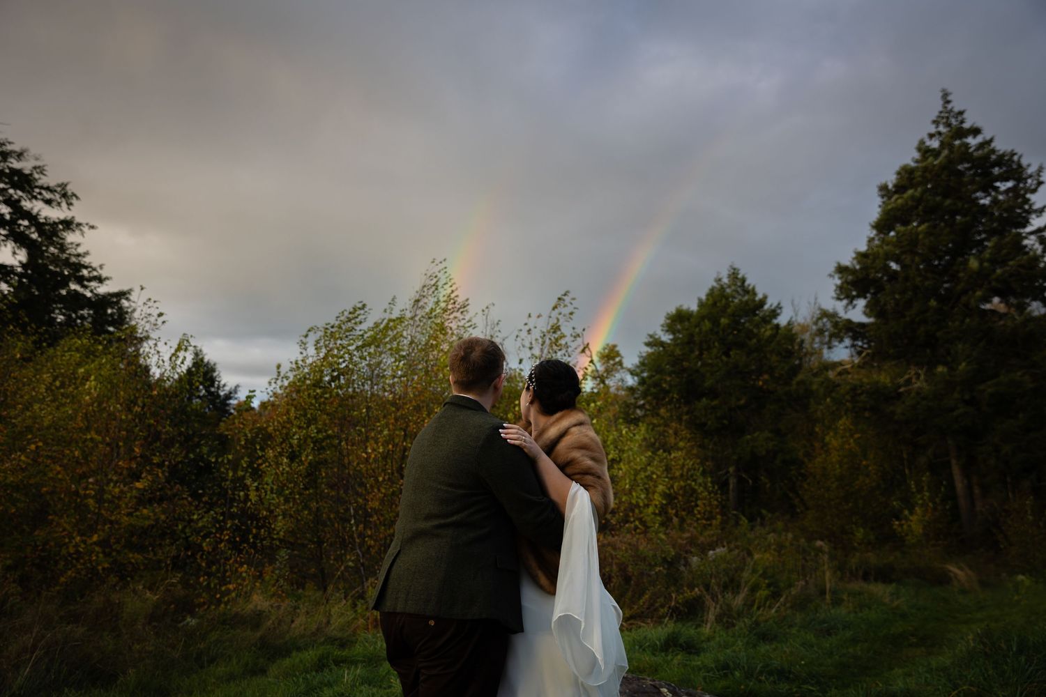 Newlyweds embrace while two vibrant rainbows appear in the cloudy sky behind them.