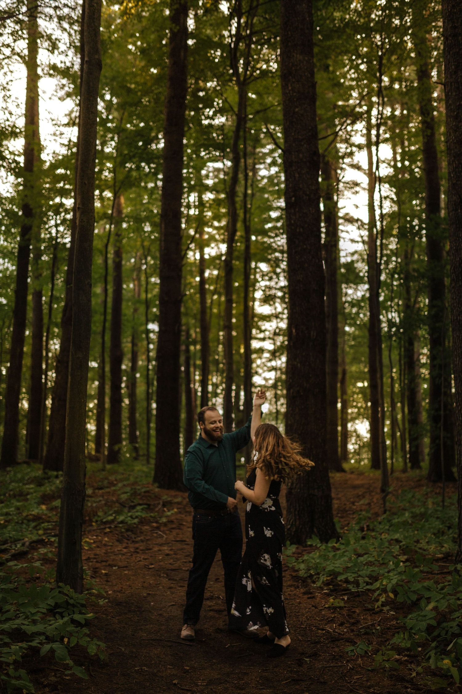 A couple walks intimately through a tall evergreen forest on a nature trail.