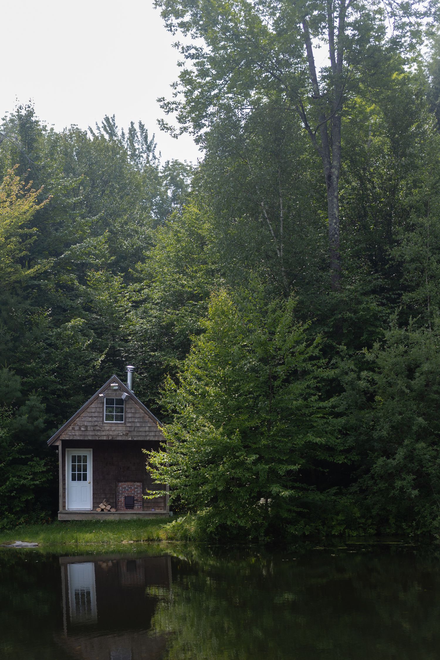 Small rustic cabin reflected in a calm pond surrounded by dense forest.