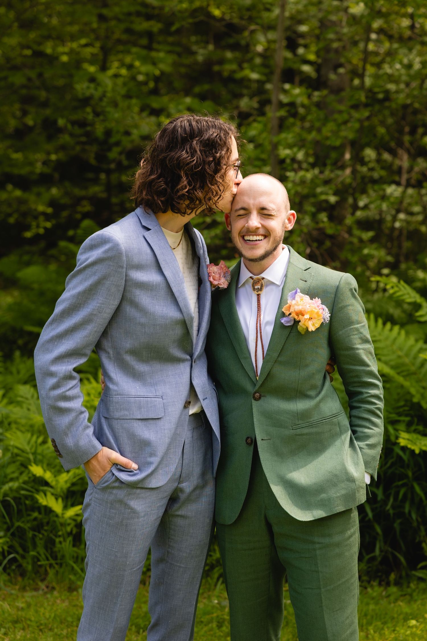Two people in formal suits embrace and laugh together against a backdrop of green foliage.