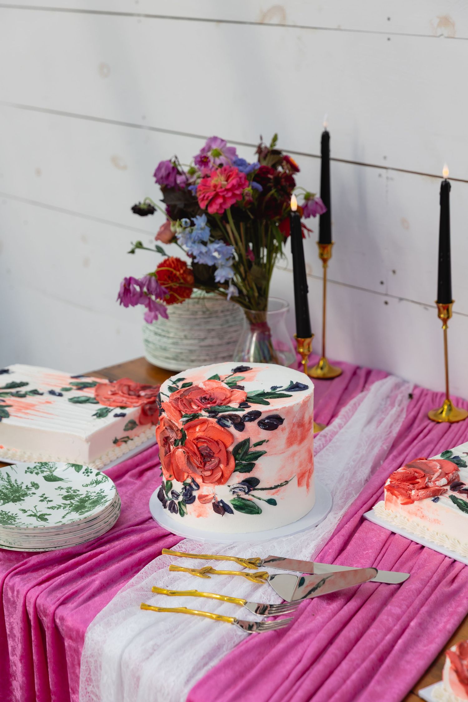 A beautifully decorated cake table with floral painted cakes, pink tablecloth, and fresh flower centerpiece.