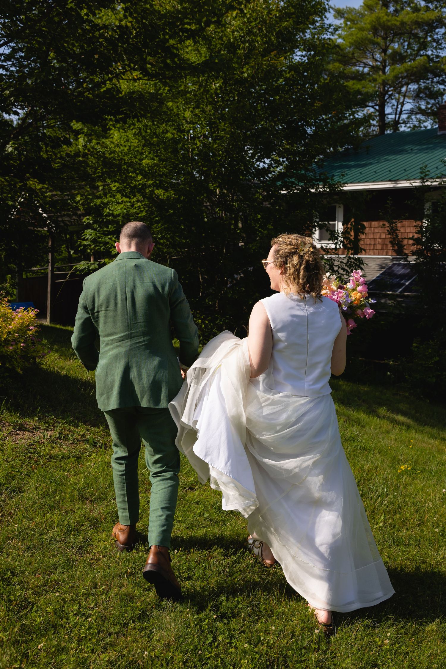 A couple in wedding attire walks through a grassy hillside path surrounded by trees on a sunny day.