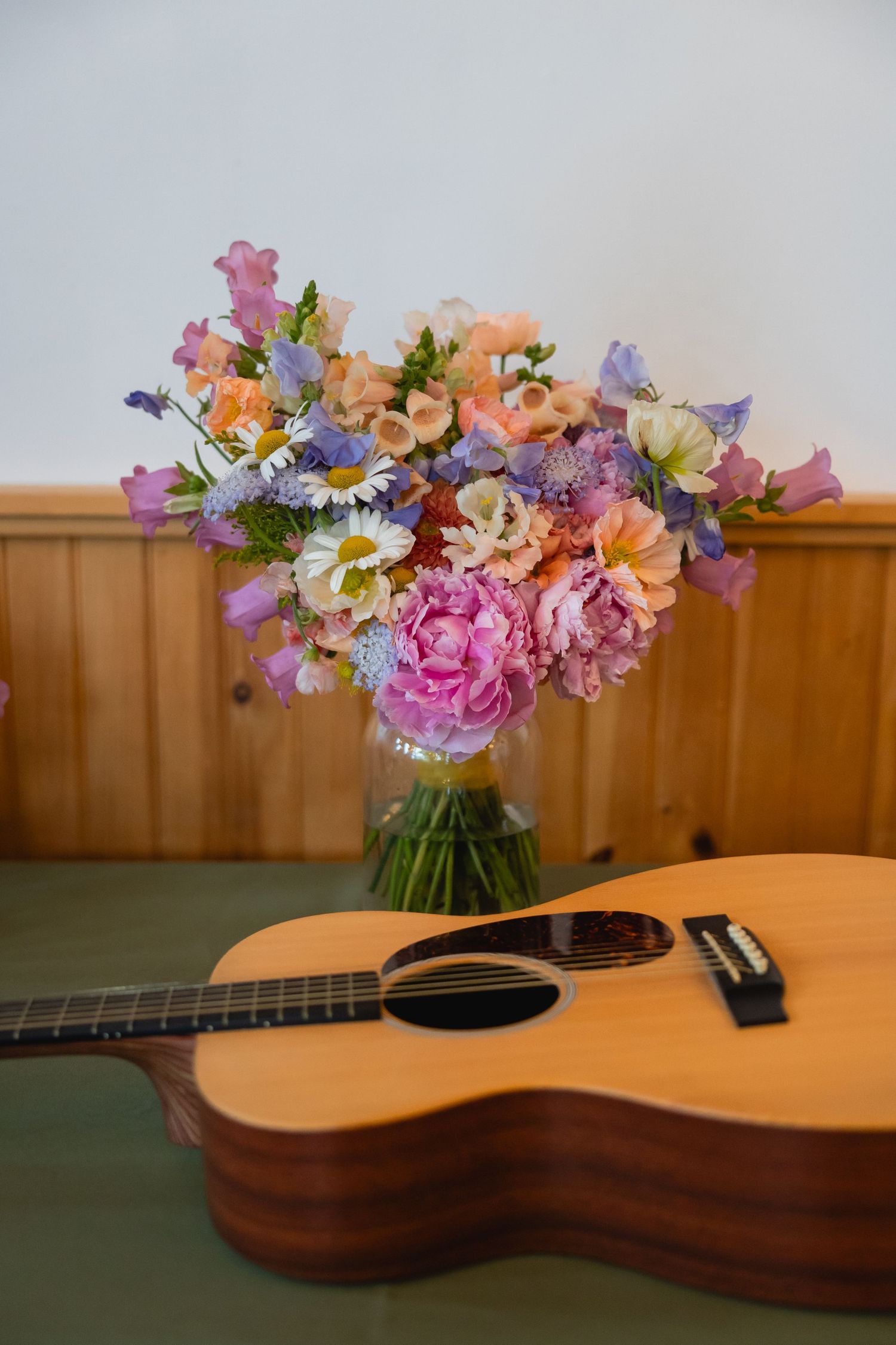 A beautiful floral arrangement of pink peonies and mixed flowers sits next to a wooden acoustic guitar on a table.